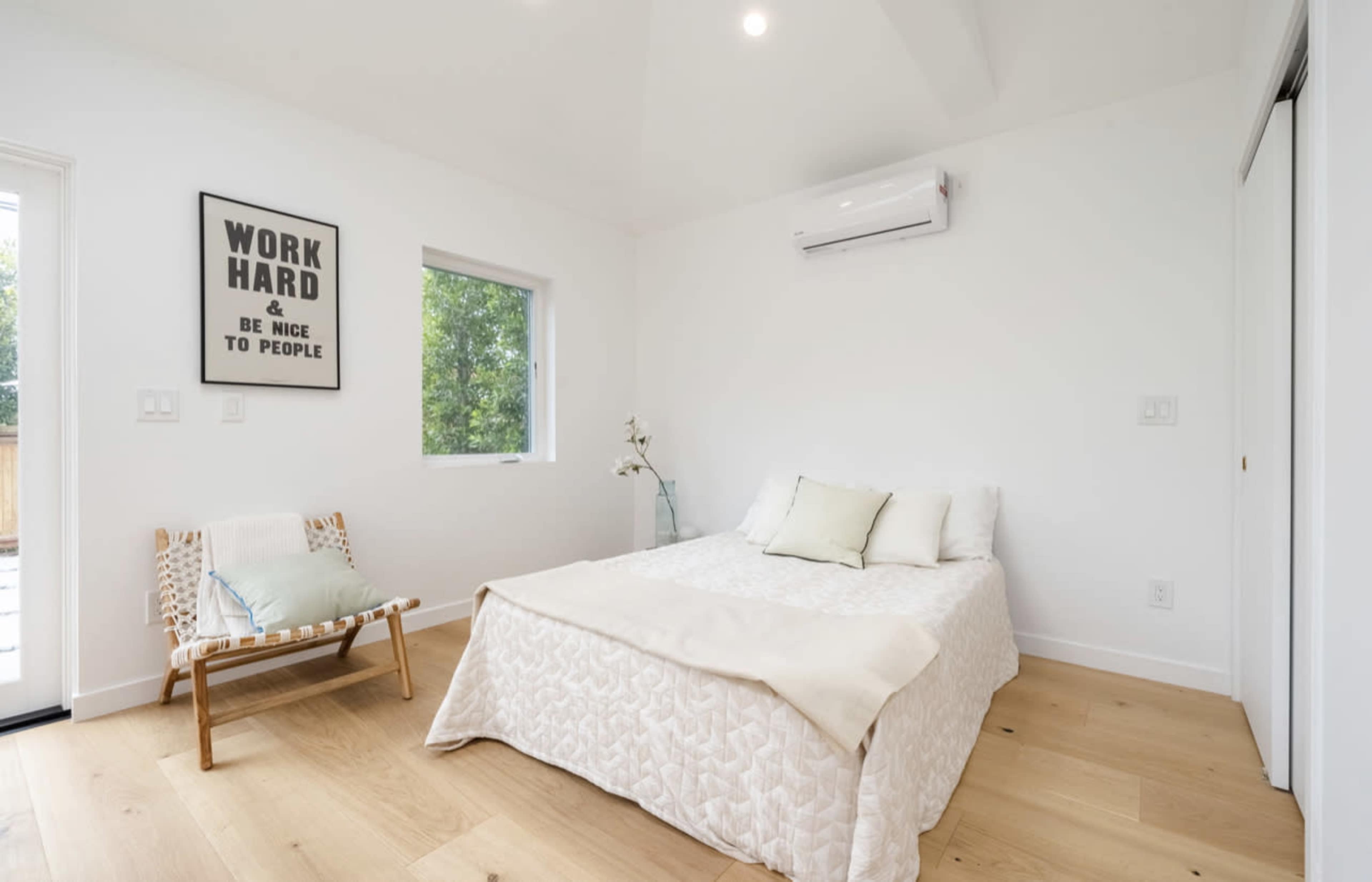 A minimalistic bedroom features a bed with light bedding, a chair in the corner, and a large window allowing natural light.