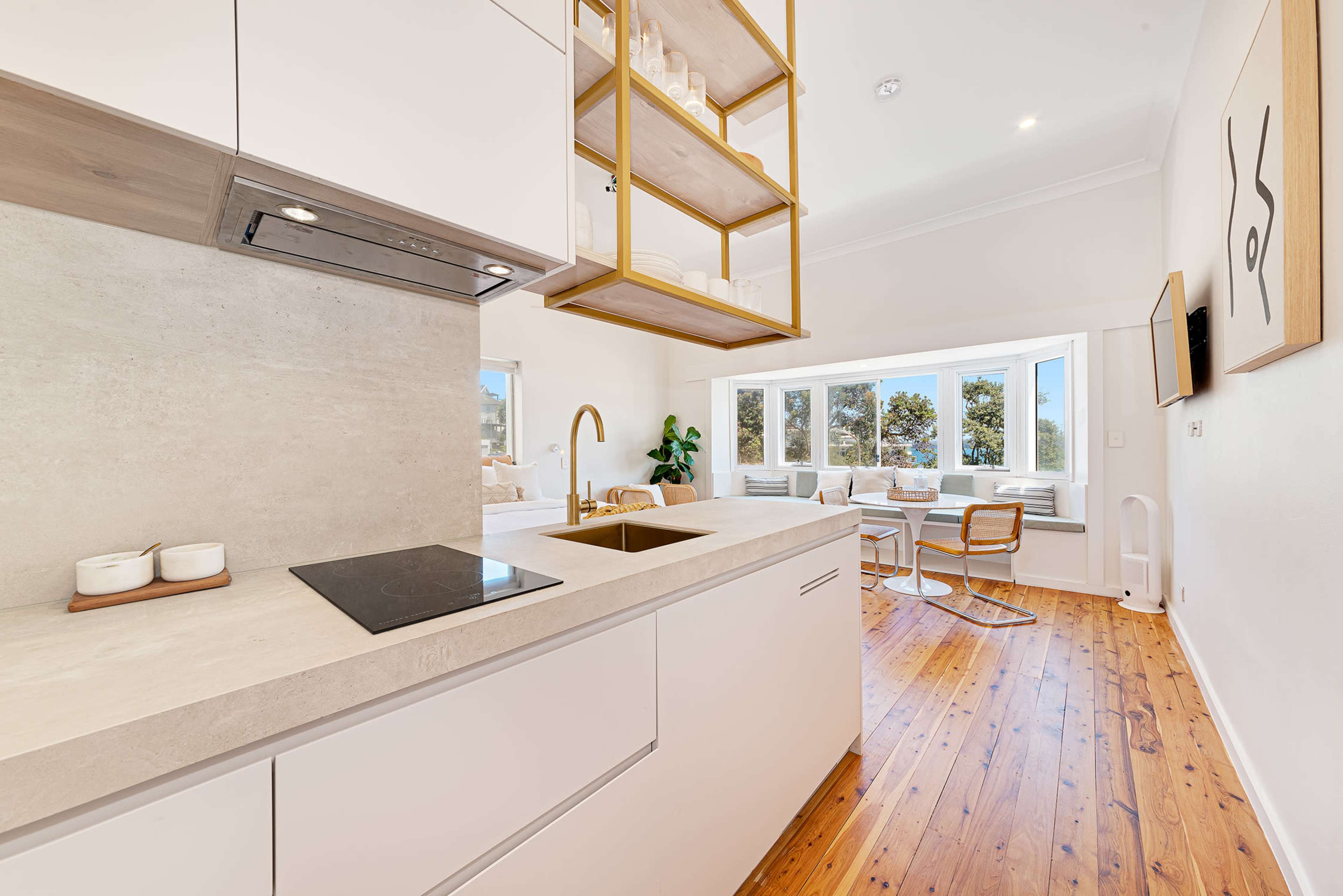 A modern kitchen features light-colored cabinetry, a sleek countertop, and a hanging display shelf, with a view of a bright dining area in the background.