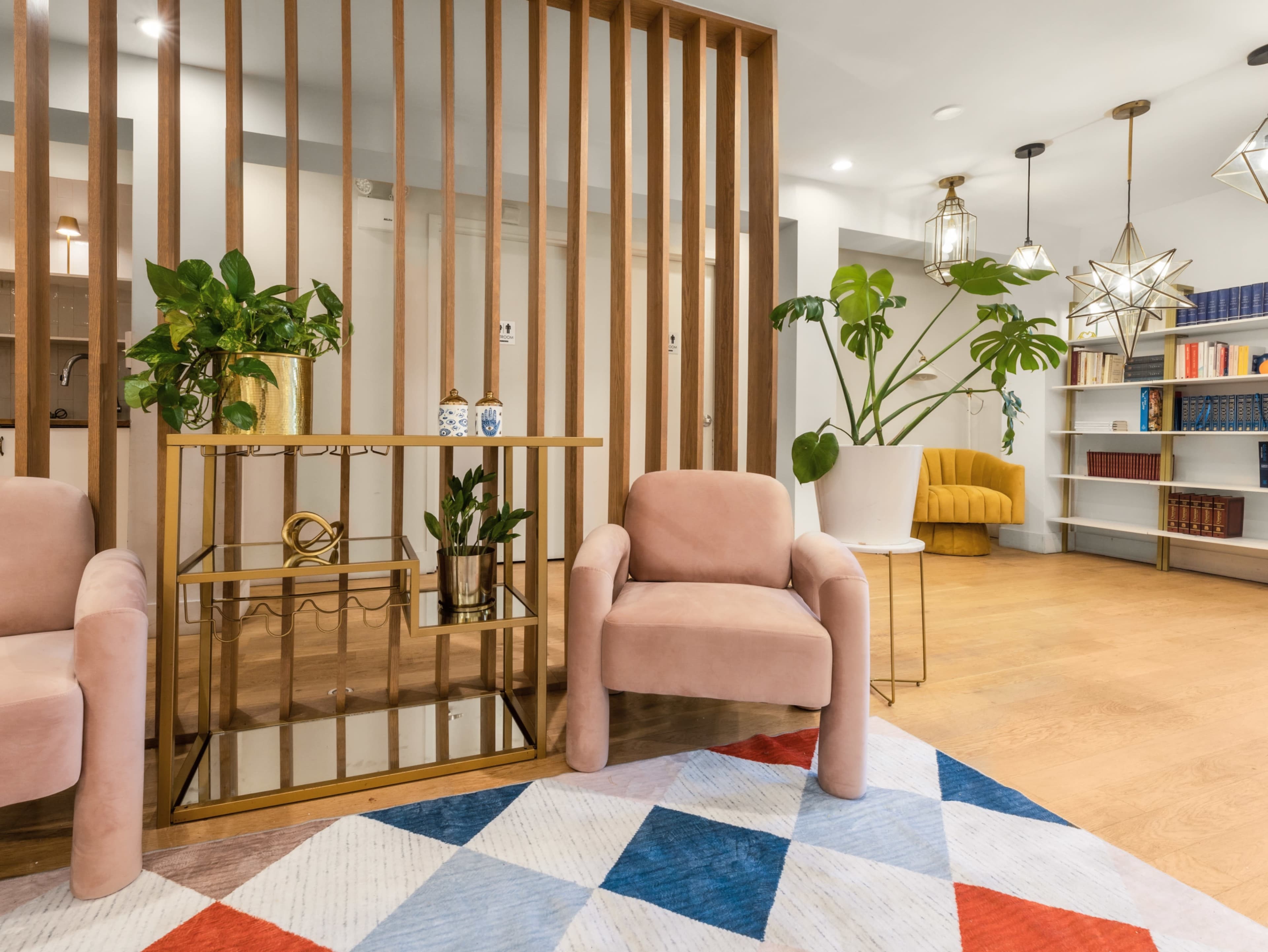 The image shows a modern lobby area with two pink chairs, a geometric rug, decorative plants, and a gold-accented shelf against a backdrop of wooden slats and pendant lights.