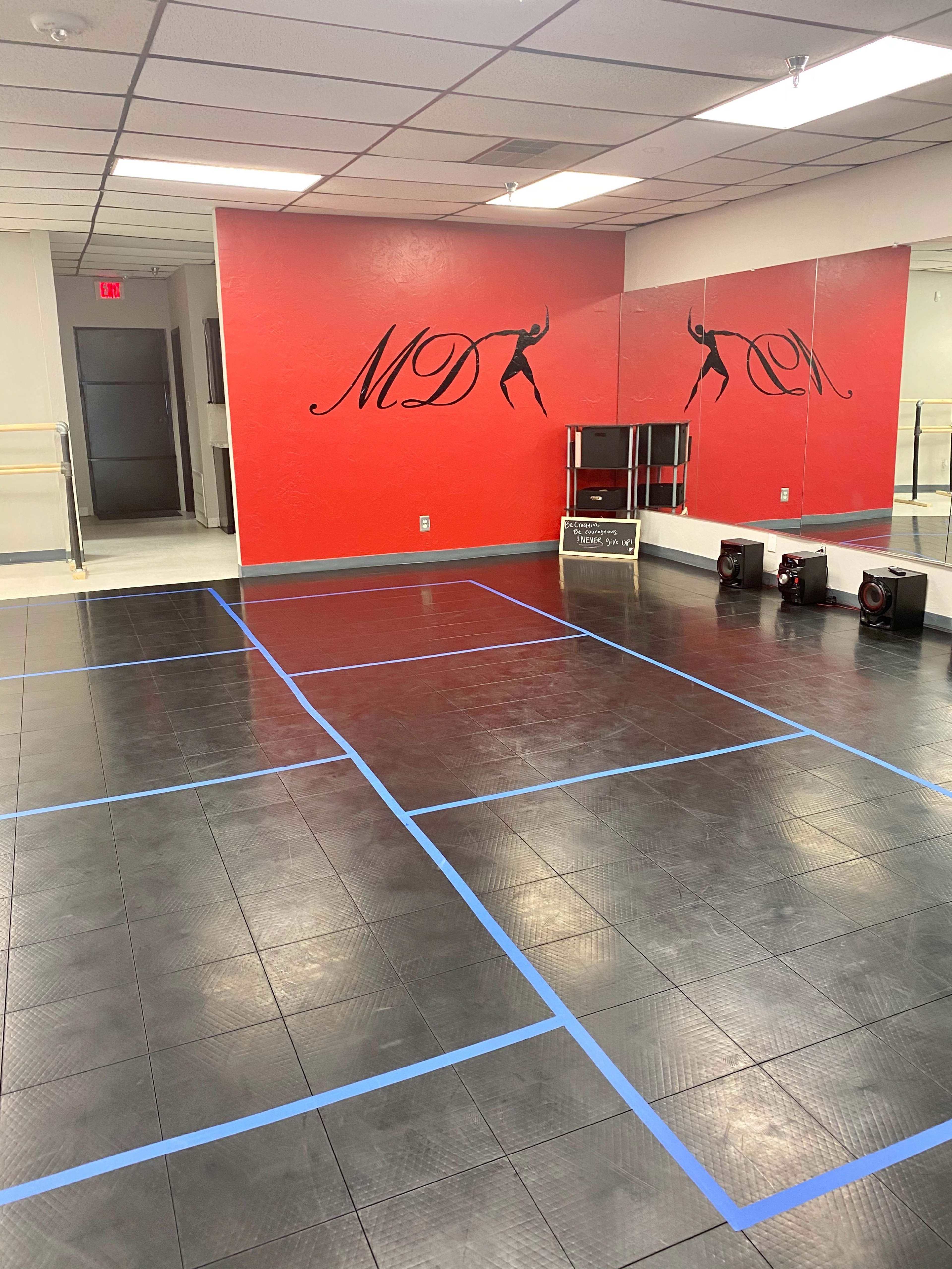 A dance studio with a mirrored wall, red accents, and a floor marked by blue tape in a grid pattern.