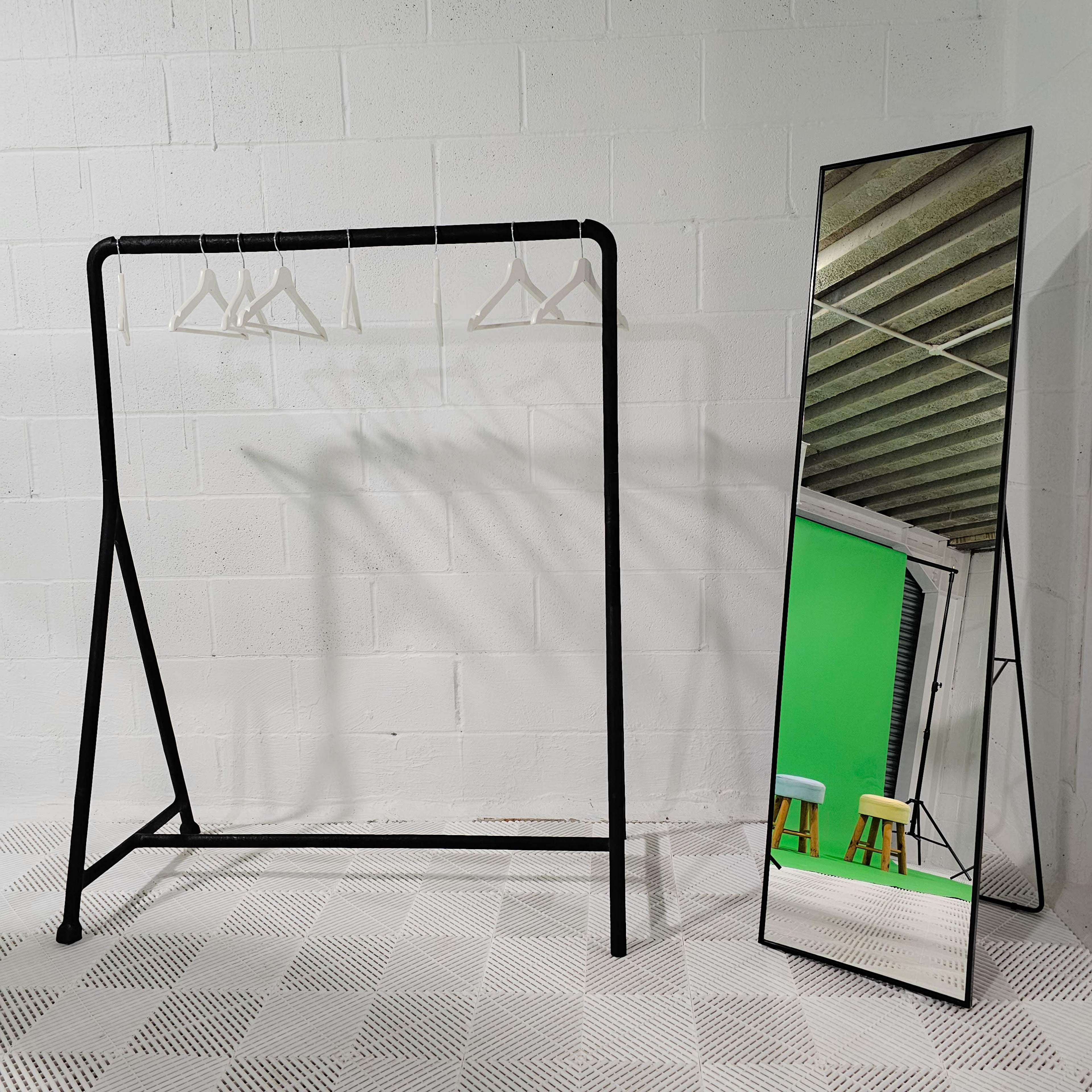 A black clothing rack with hangers stands next to a tall mirror against a white wall, with a green backdrop visible behind the mirror.