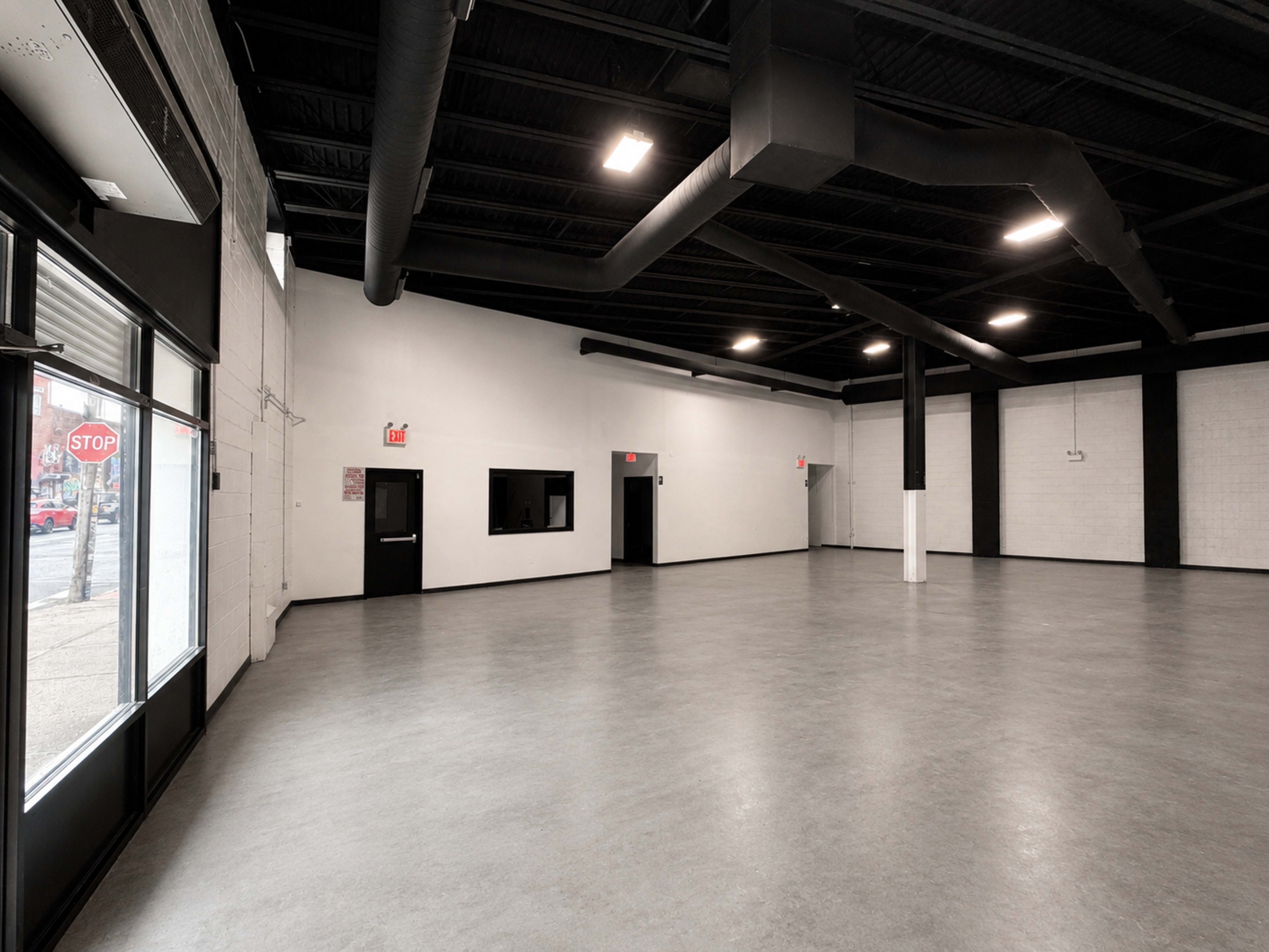 The image shows a spacious, empty room with gray flooring, high black ceilings, and large windows letting in natural light.