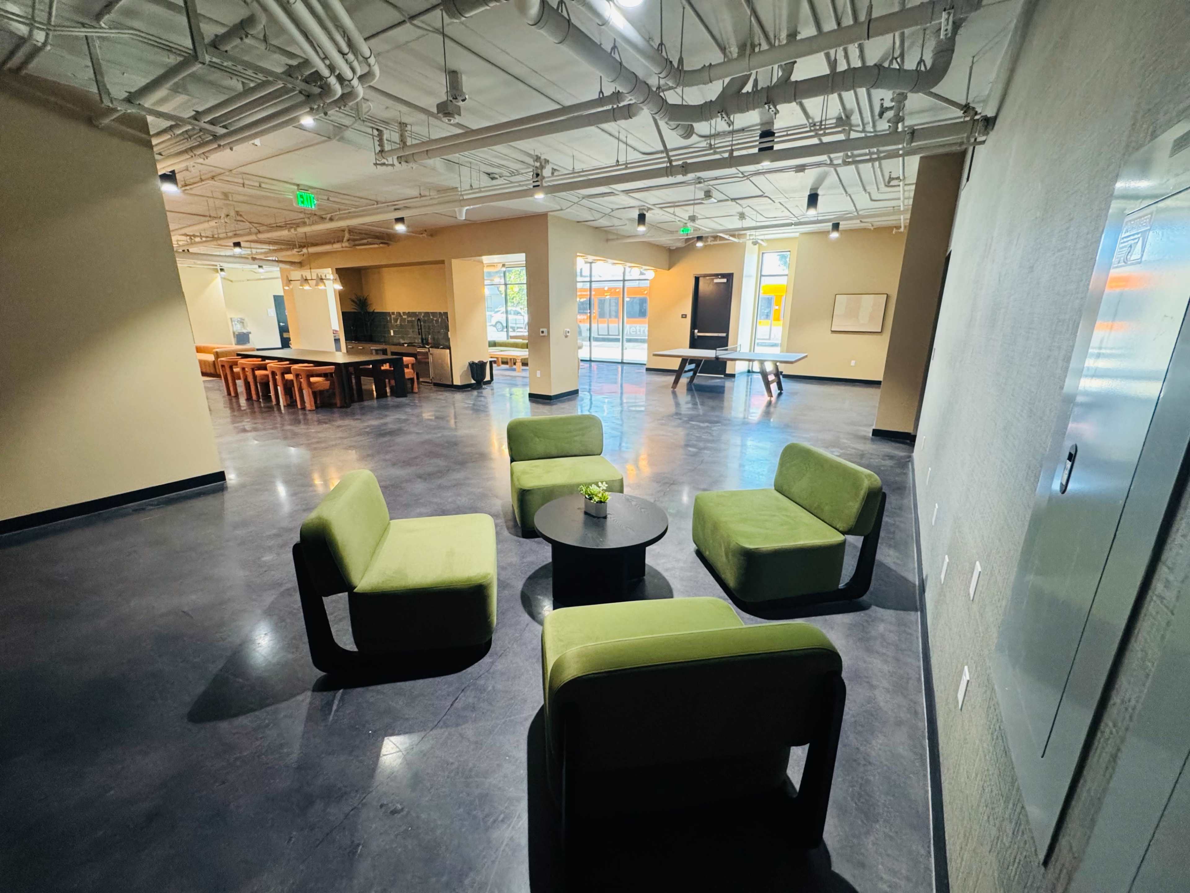 The image shows a modern, open space featuring four green armchairs arranged around a small black table, with a communal dining area and a ping pong table in the background.