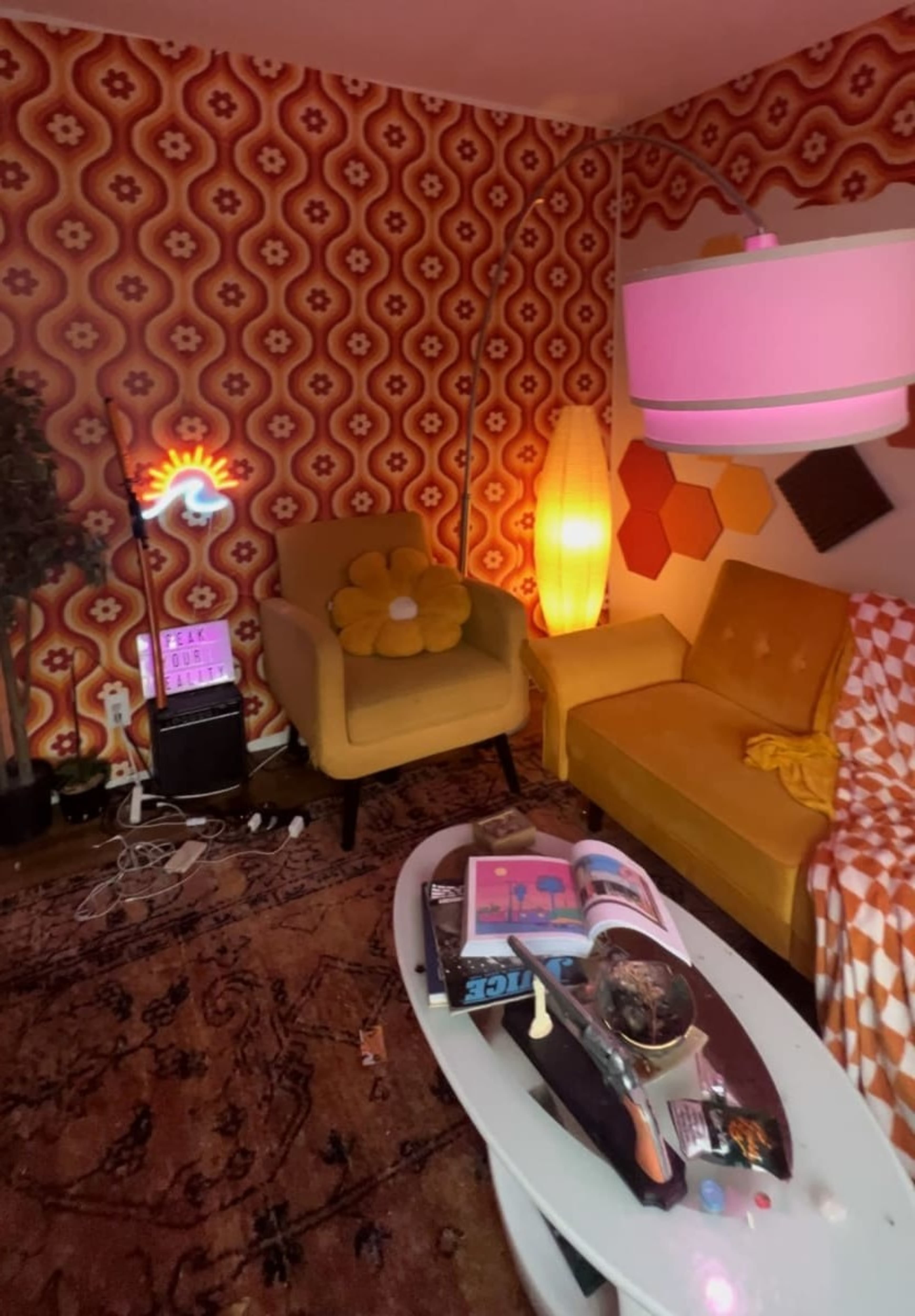 A retro-styled living room with patterned wallpaper, a yellow sofa, an armchair with a flower design, a coffee table cluttered with books and decorative items, and various lighting sources.