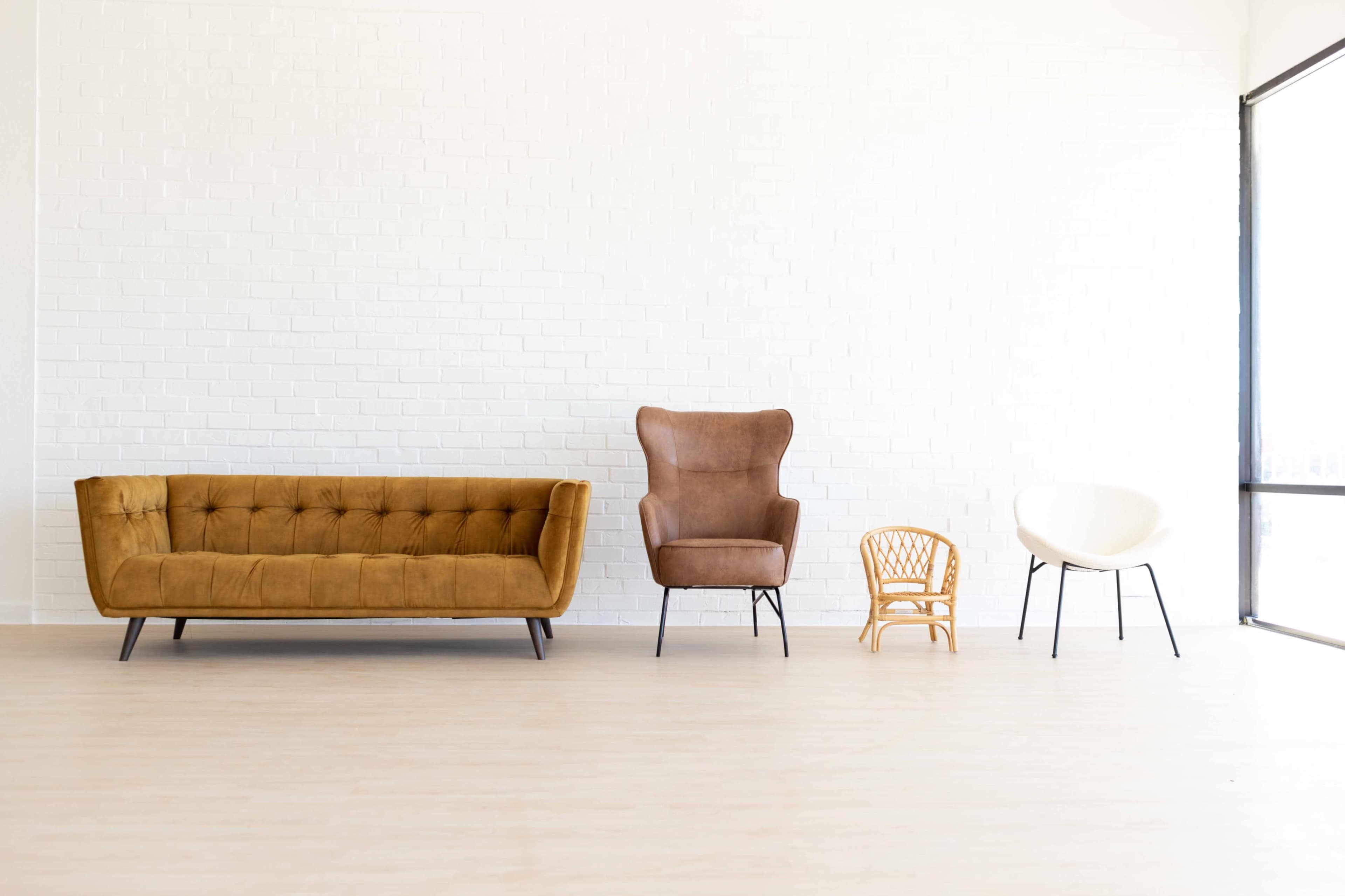A minimalist interior featuring a brown velvet sofa, a brown upholstered armchair, a small wooden chair, and a white circular chair against a white brick wall.