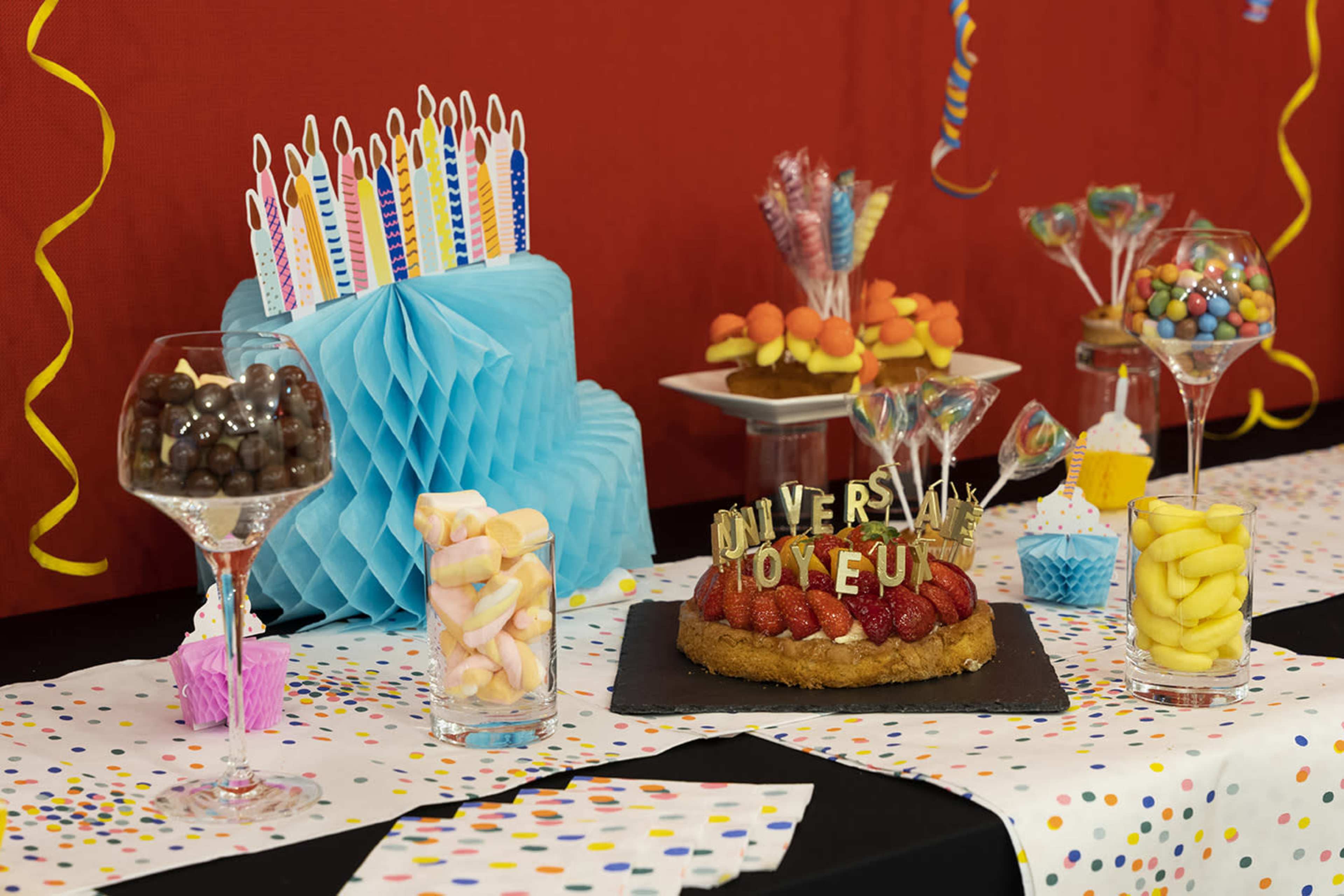 A colorful birthday party table displays a cake with decorative candles, candy-filled glasses, and various sweets arranged on a polka-dotted tablecloth.
