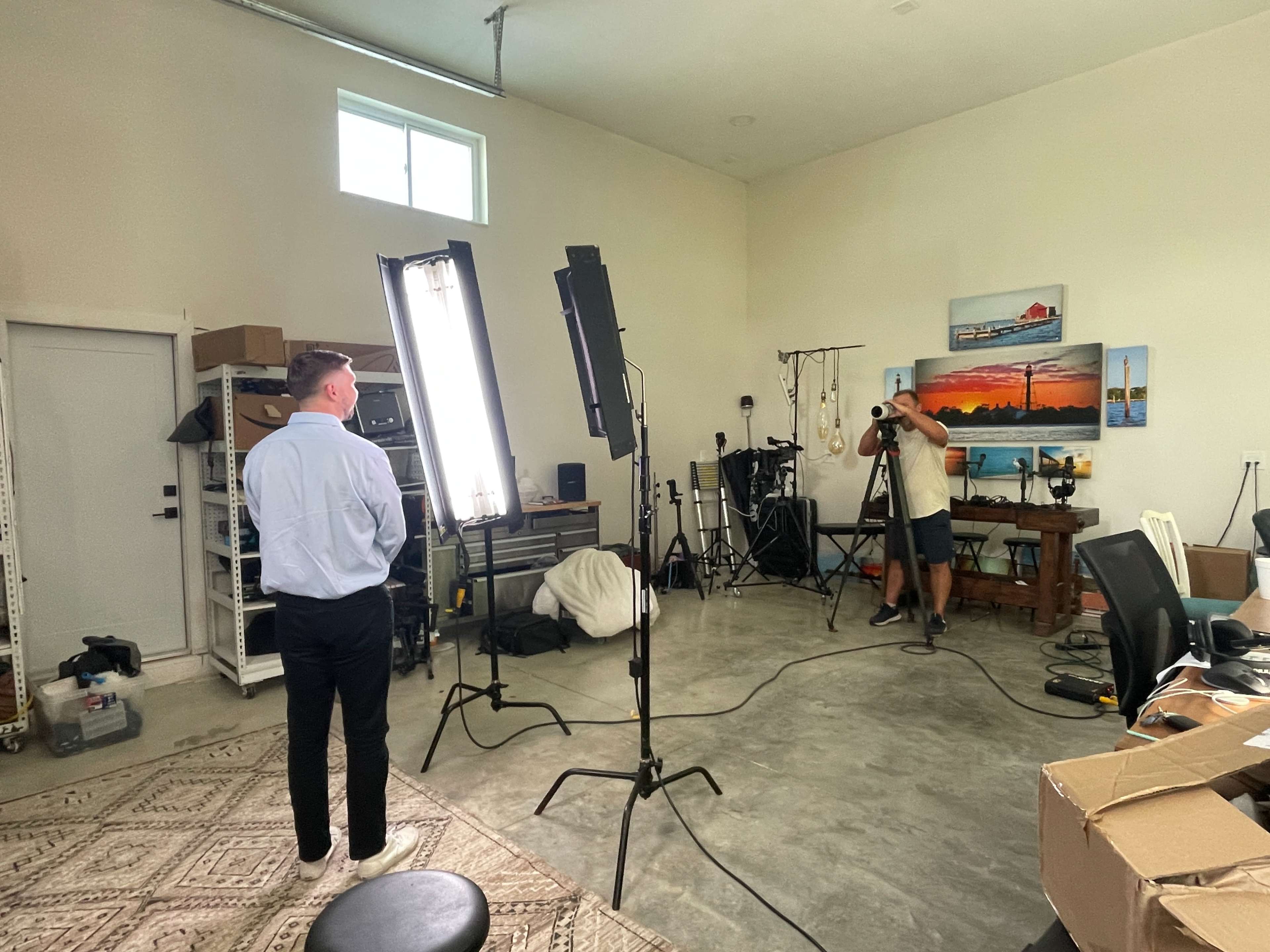 A person in a light blue shirt stands in front of two bright lights while another person holds a camera in a studio-like setting.
