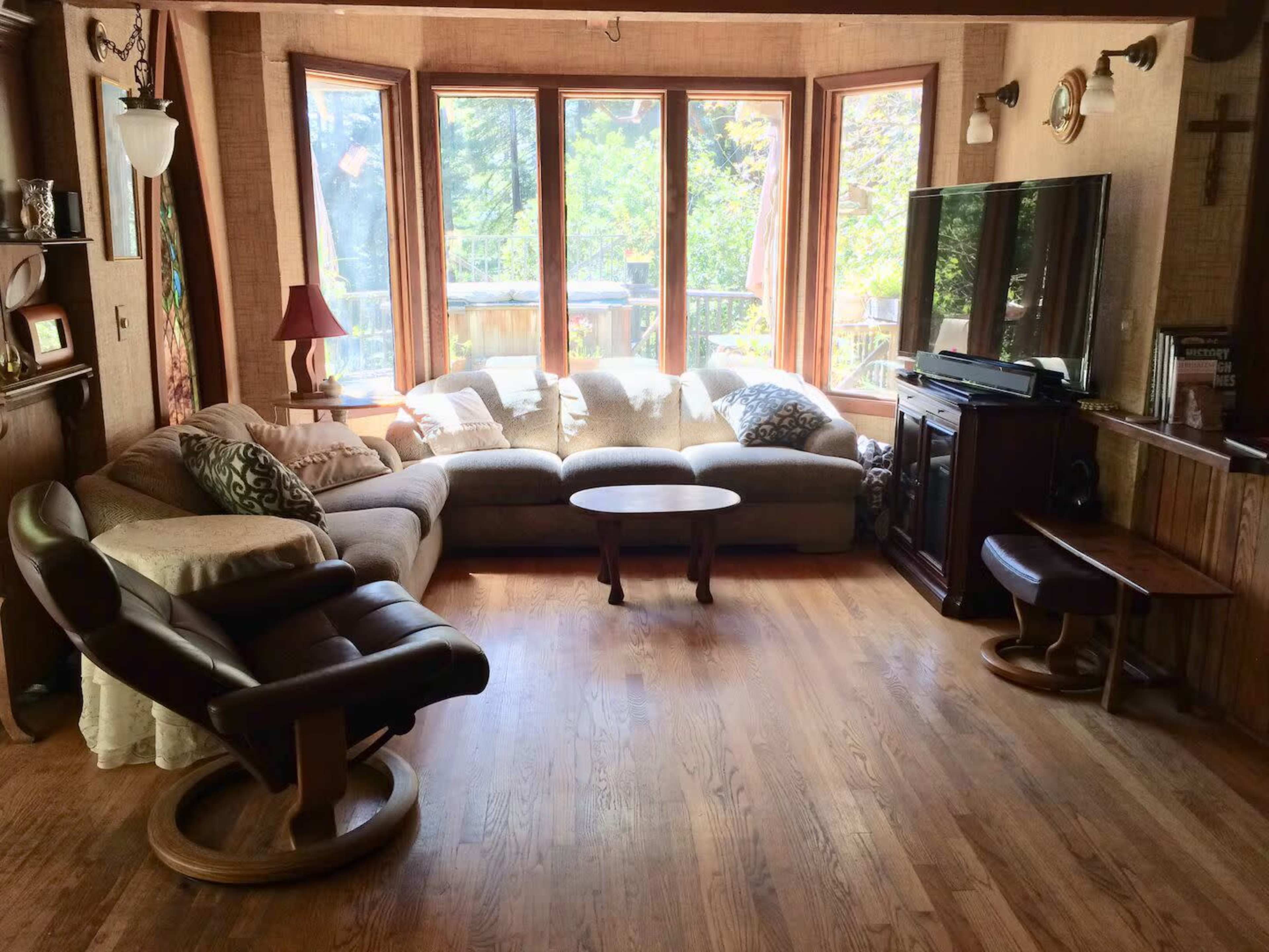 The image shows a cozy living room with a large sectional sofa, a coffee table, and a flat-screen TV, all surrounded by large windows letting in natural light.