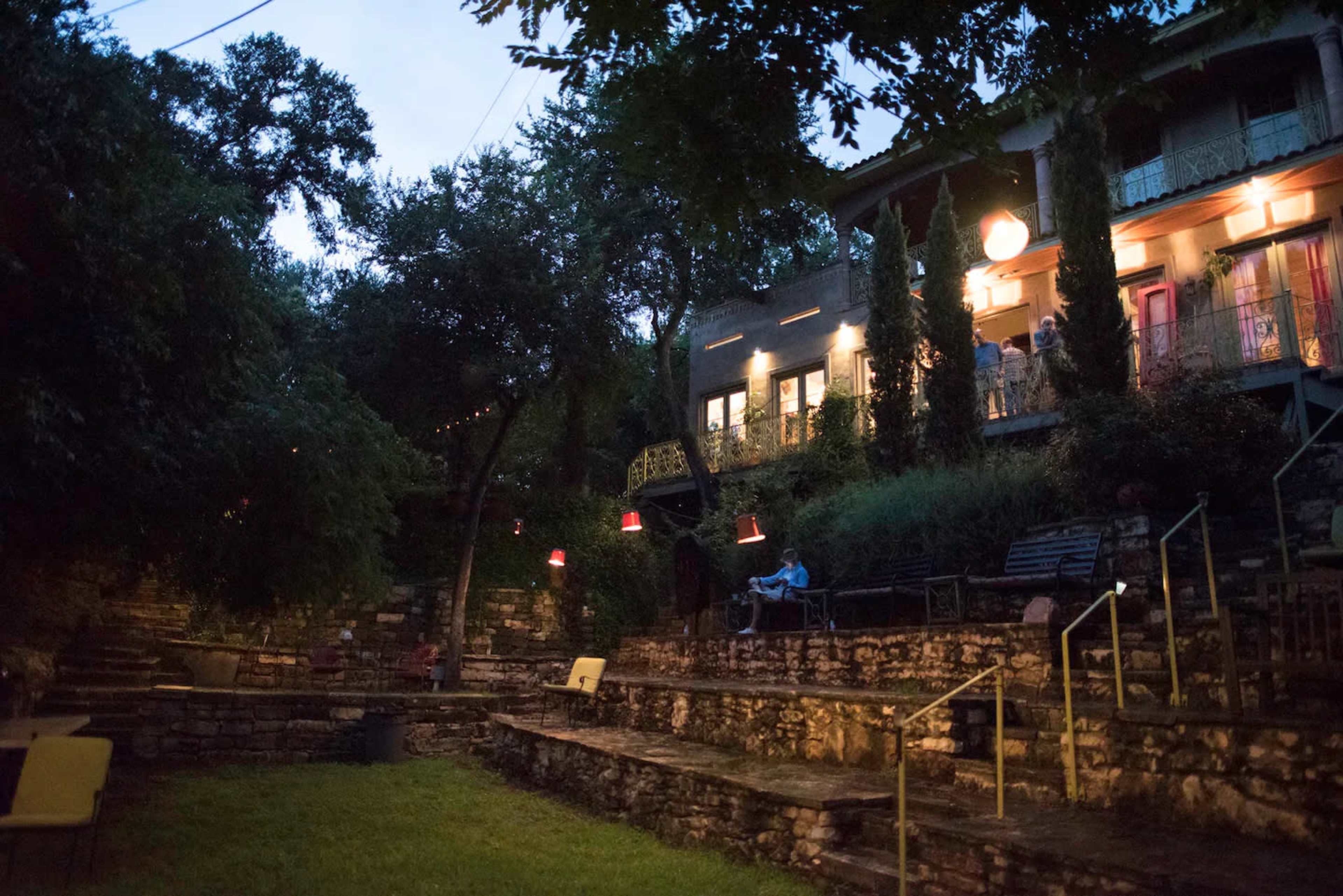 The image shows a dimly lit hillside garden area with stone steps leading up to a multi-level building, surrounded by trees and decorative lights.