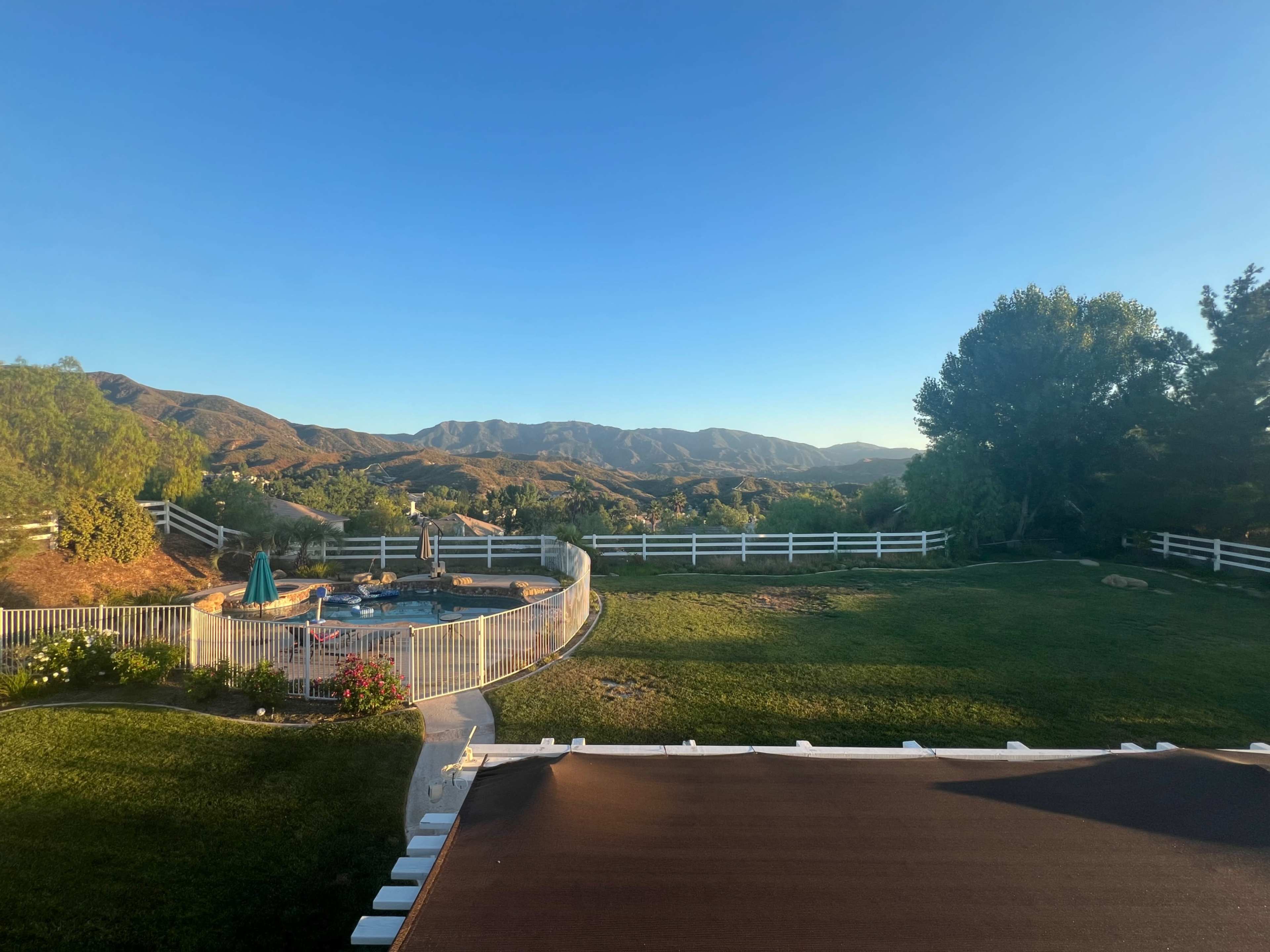 The image shows a backyard with a swimming pool surrounded by a fence, set against a backdrop of rolling hills and clear blue skies.