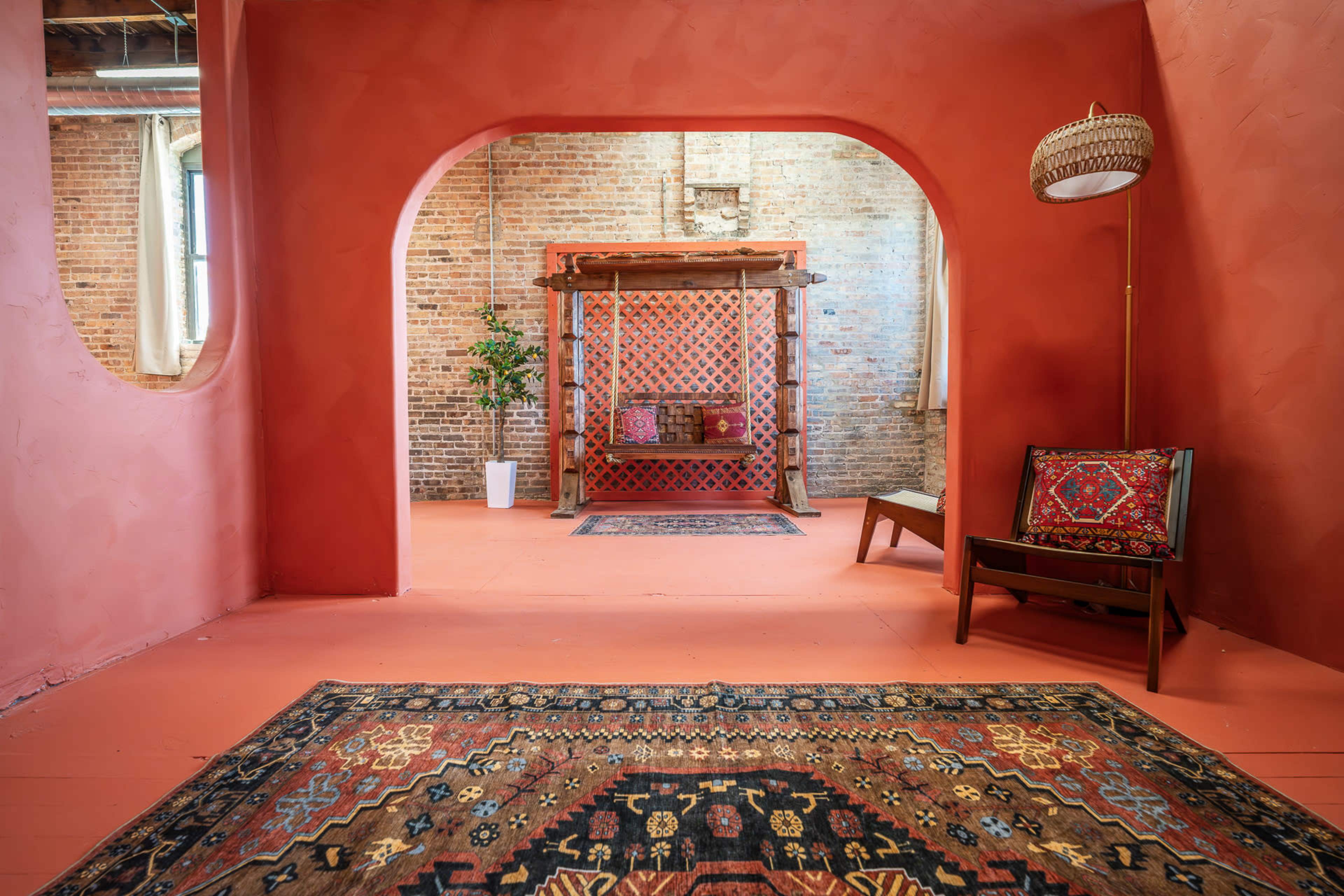 The image depicts a room with red walls, a wooden structure in the center adorned with latticework, and decorative rugs on the floor, complemented by a plant and a floor lamp.