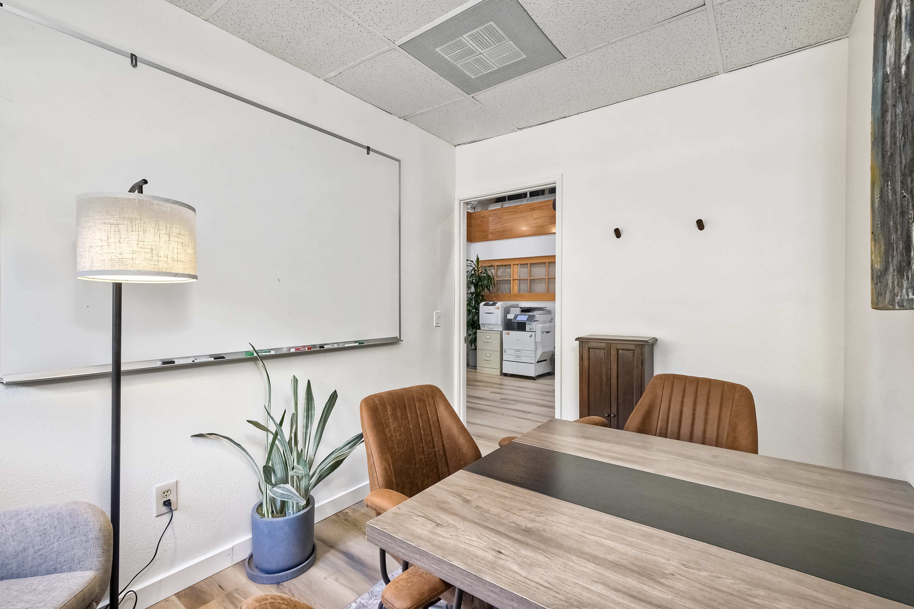 The image shows a small meeting room with a wooden table, four chairs, a whiteboard, a lamp, a plant, and a doorway leading to another room.