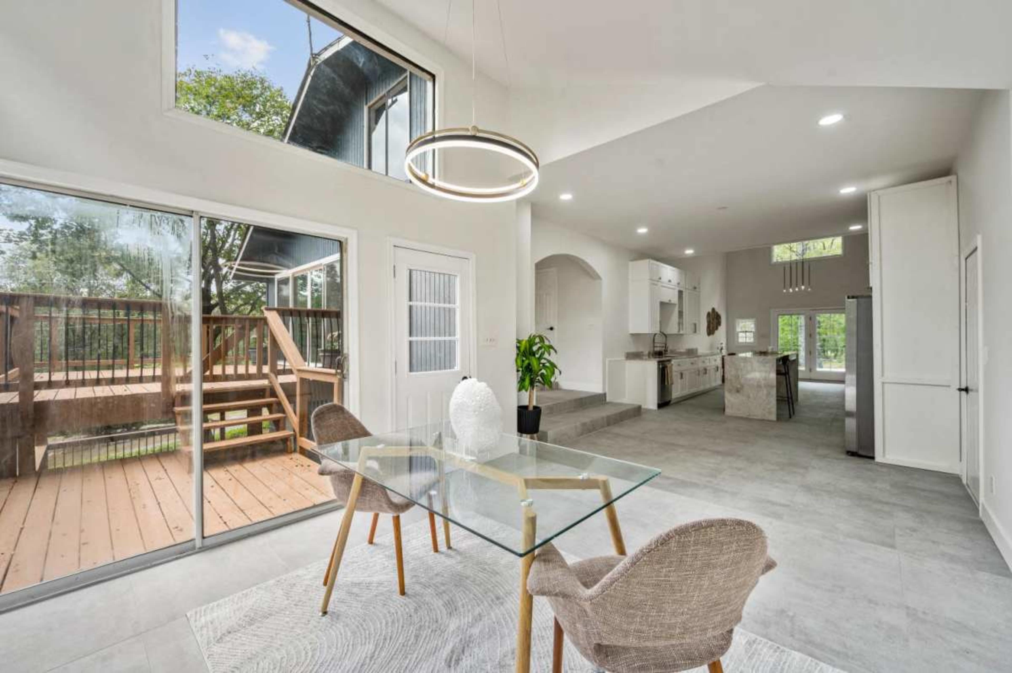 The image shows a modern dining area with a glass table and two chairs, overlooking a wooden deck through large sliding glass doors.