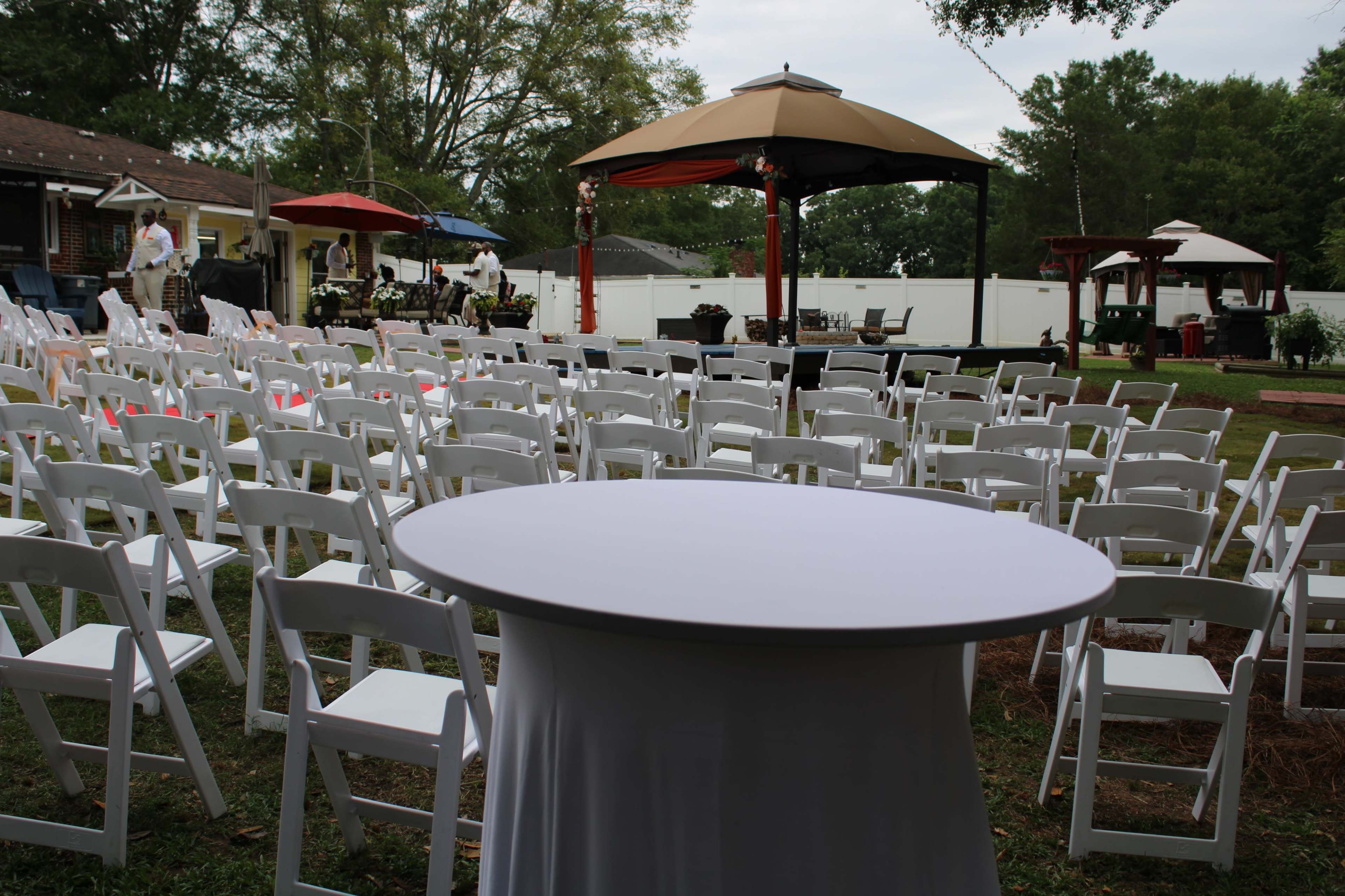 The image shows a neatly arranged outdoor event space with white chairs set up in rows, a central round table, and a large gazebo in the background.