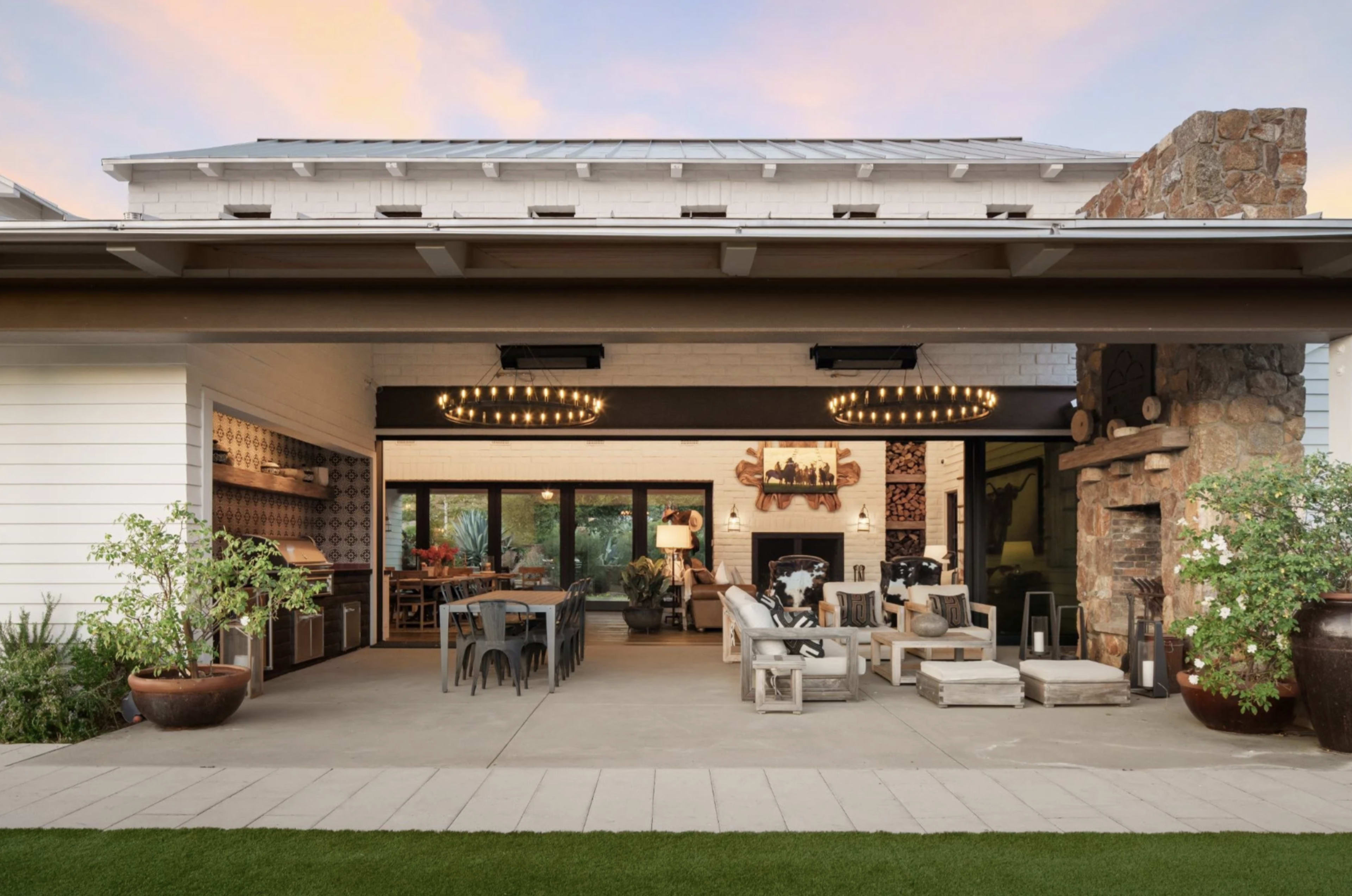 The image shows a modern outdoor living space with a spacious patio featuring a dining area, seating arrangement, and a stone fireplace, all framed by lush greenery.