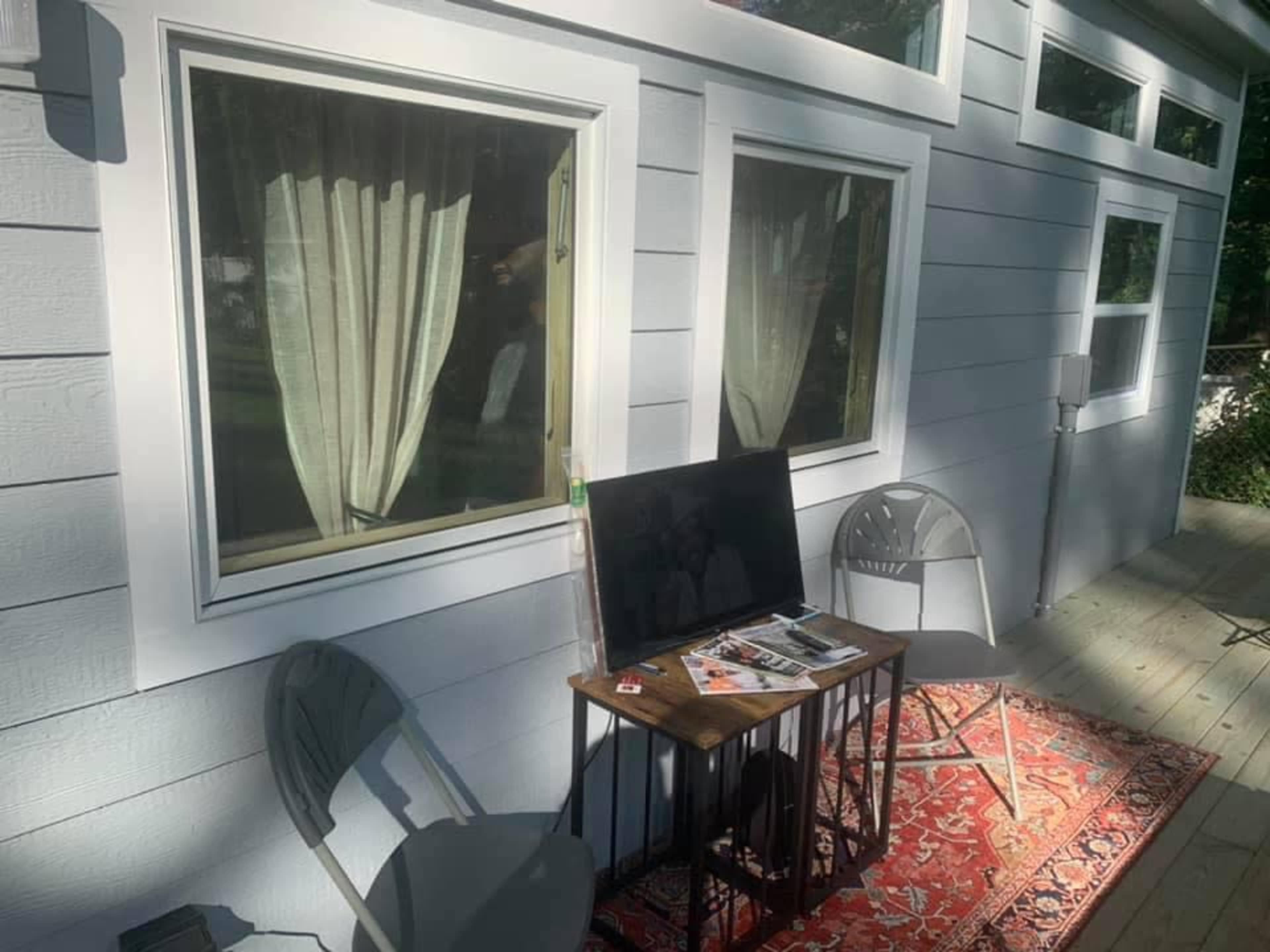 A television sits on a small table between two chairs on a porch beside a house with large windows and sheer curtains.