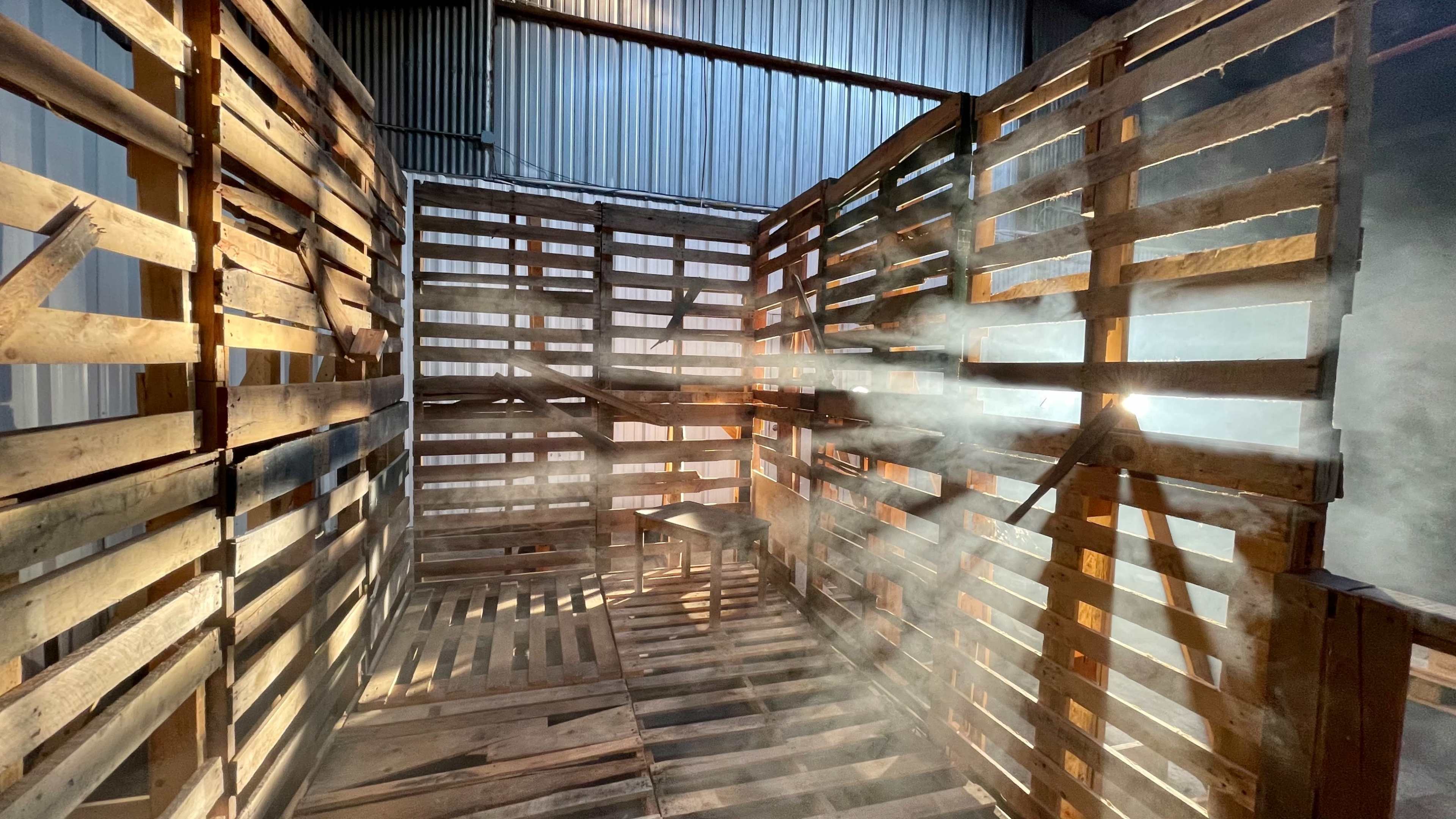 An enclosed space made of wooden pallets with beams of light piercing through gaps, creating a misty atmosphere.
