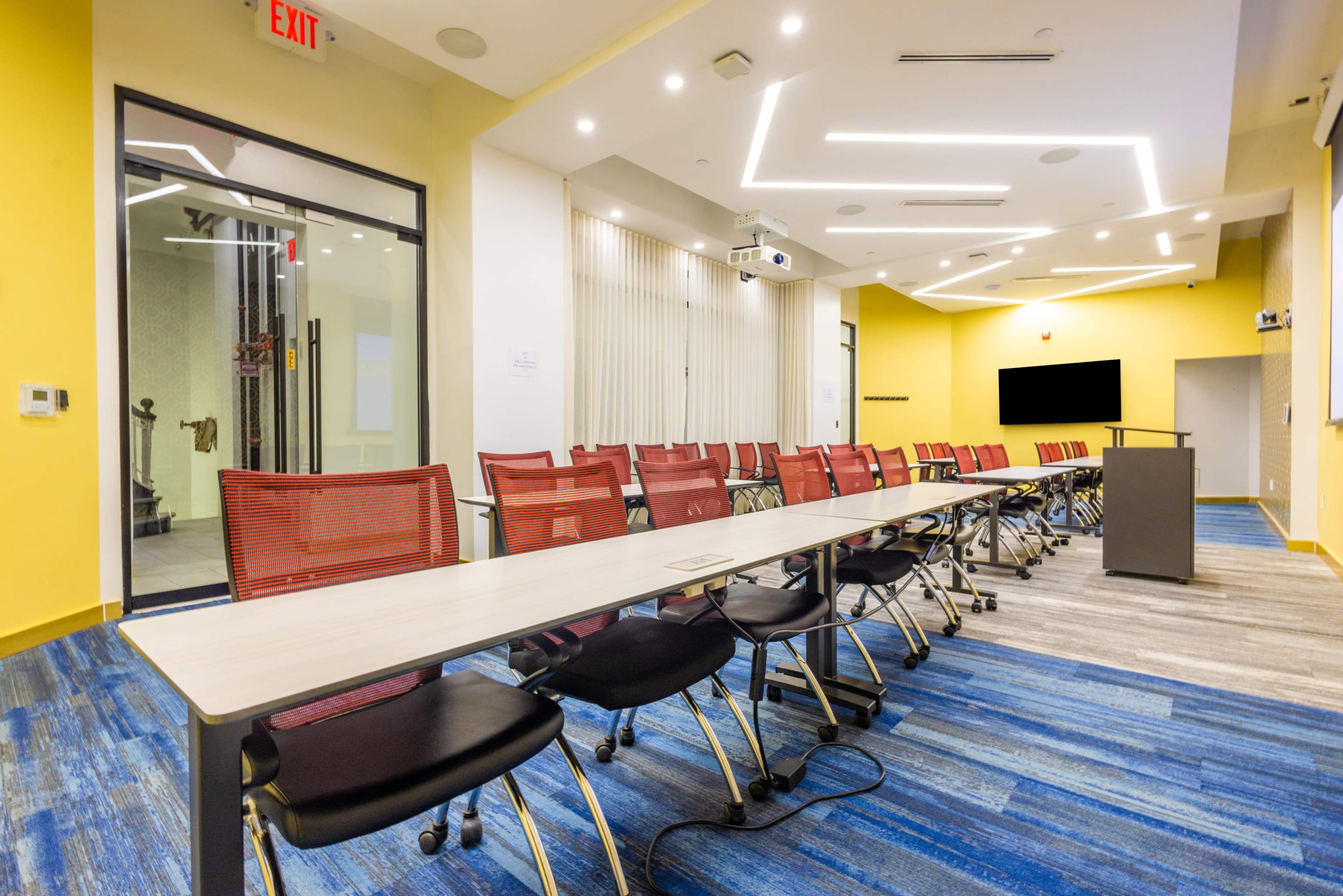 The image shows a modern conference room with a long table lined with red and black chairs, equipped with a projector and a screen at the front.