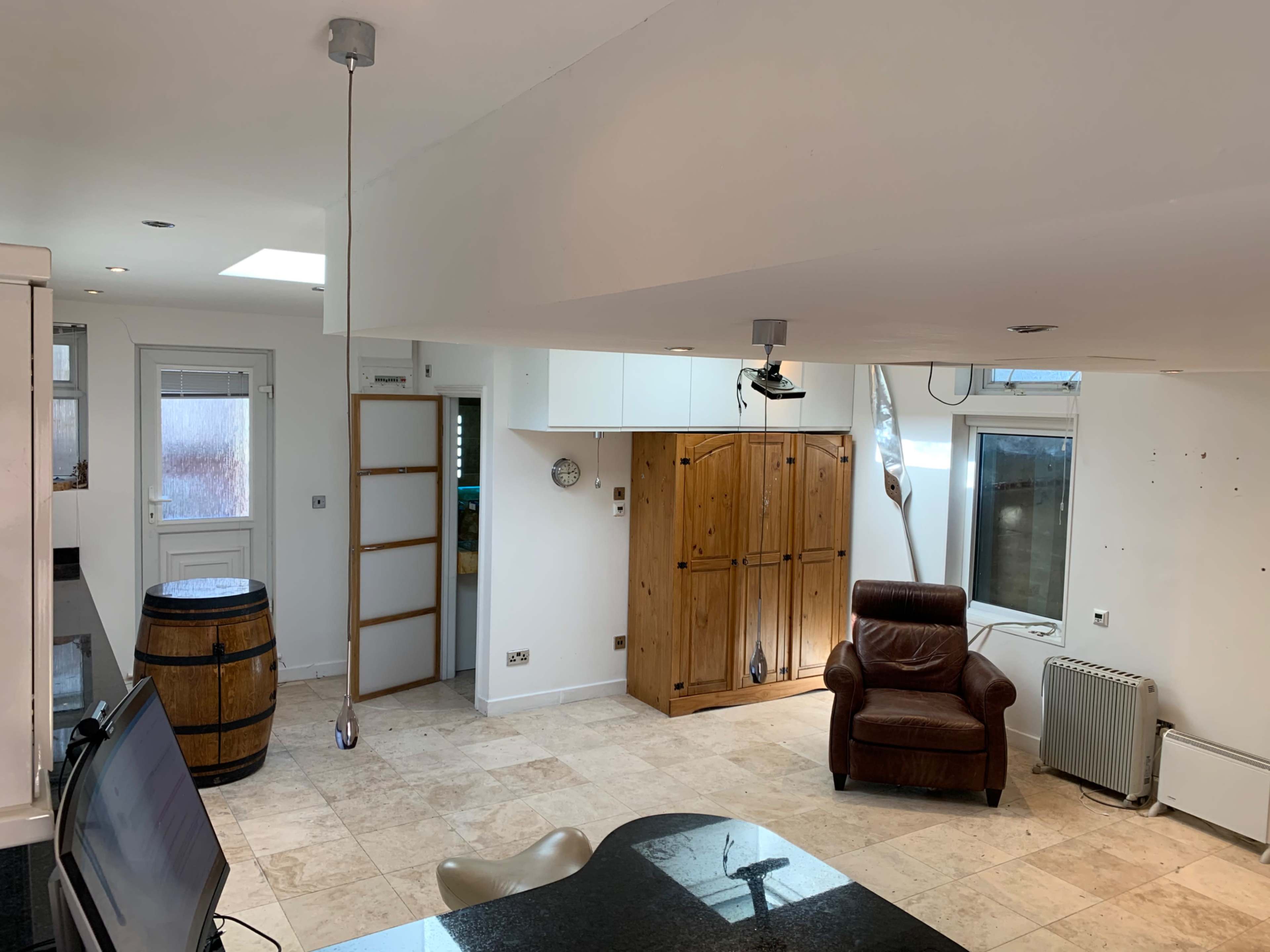 A modern, open-plan kitchen and living area with a leather armchair, a wooden cabinet, and a barrel, featuring bright white walls and tiled flooring.