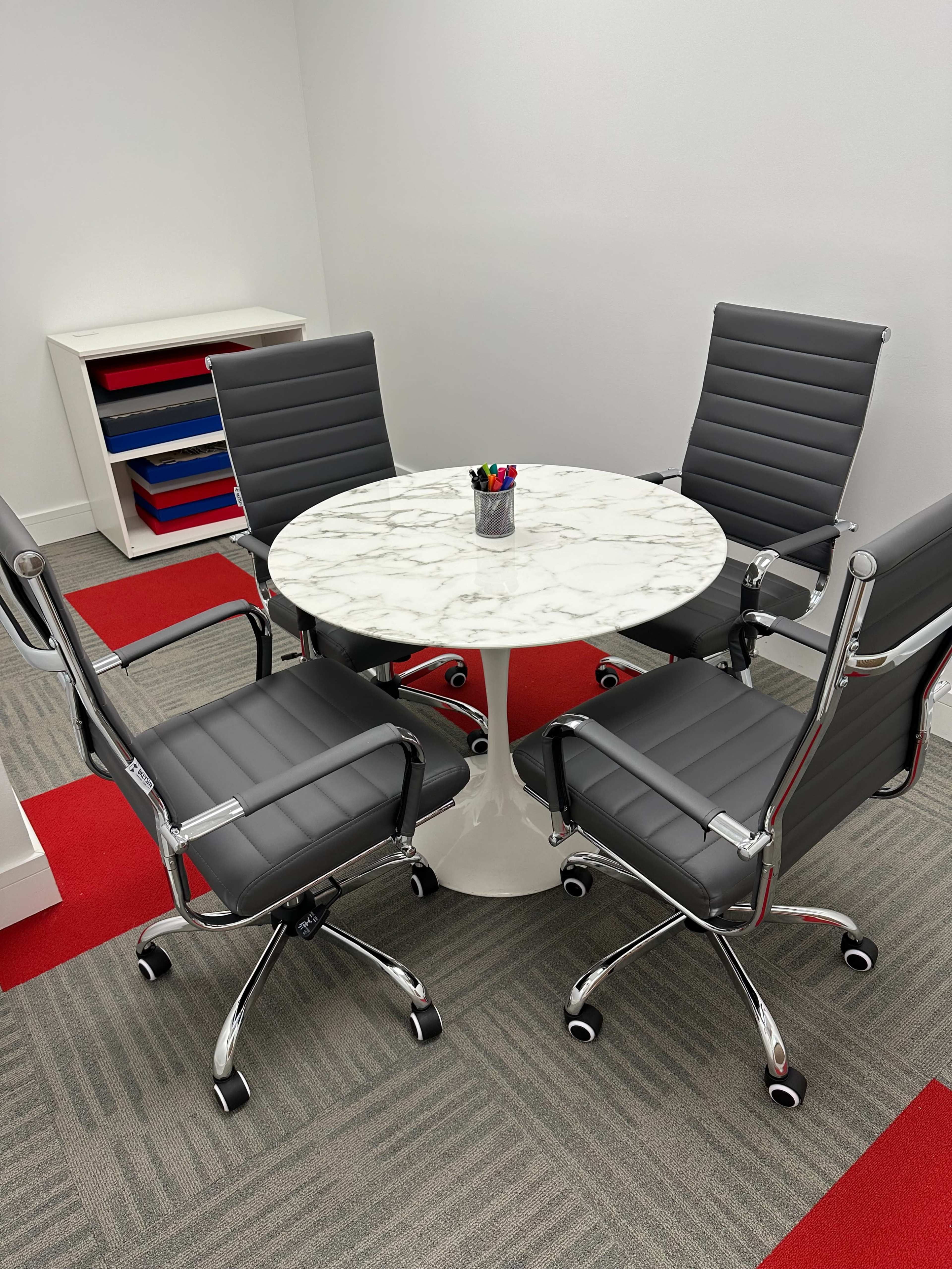 A round marble table is surrounded by four gray office chairs in a brightly colored meeting room.