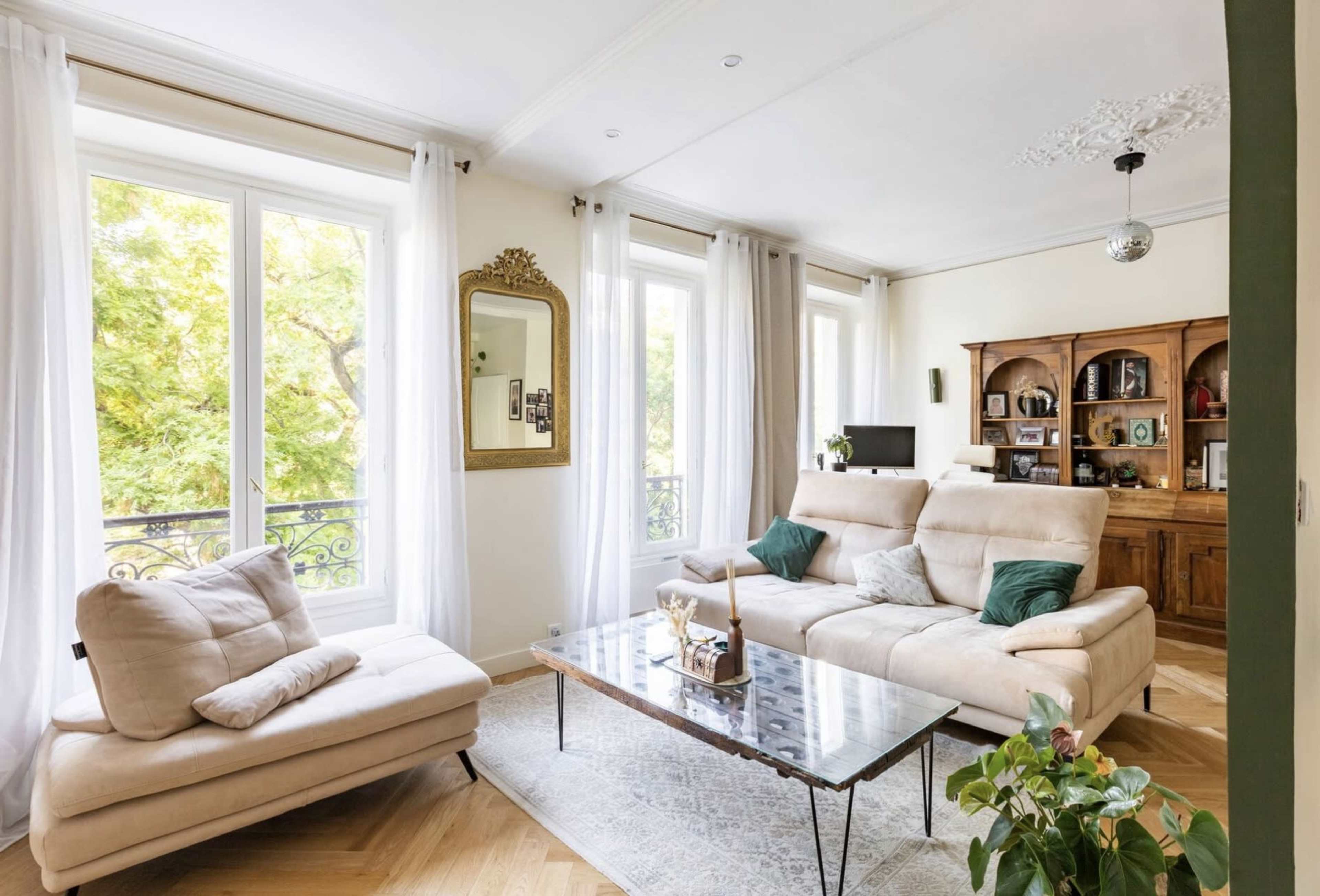 The living room features a light-colored sofa, a glass coffee table, and large windows draped with sheer curtains, allowing natural light to fill the space.