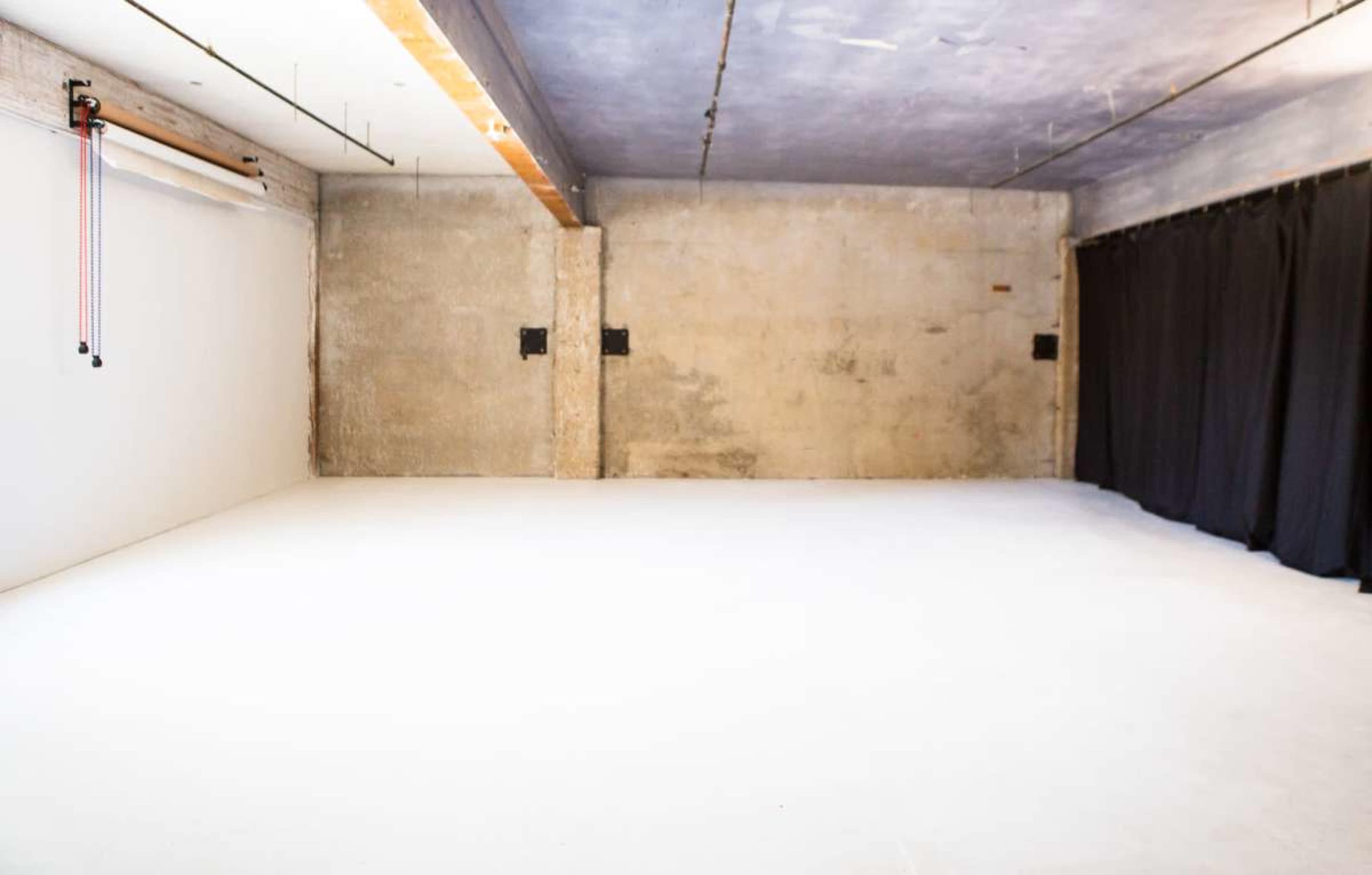 The image shows an empty studio space with a white floor, concrete walls, and black curtains on one side.