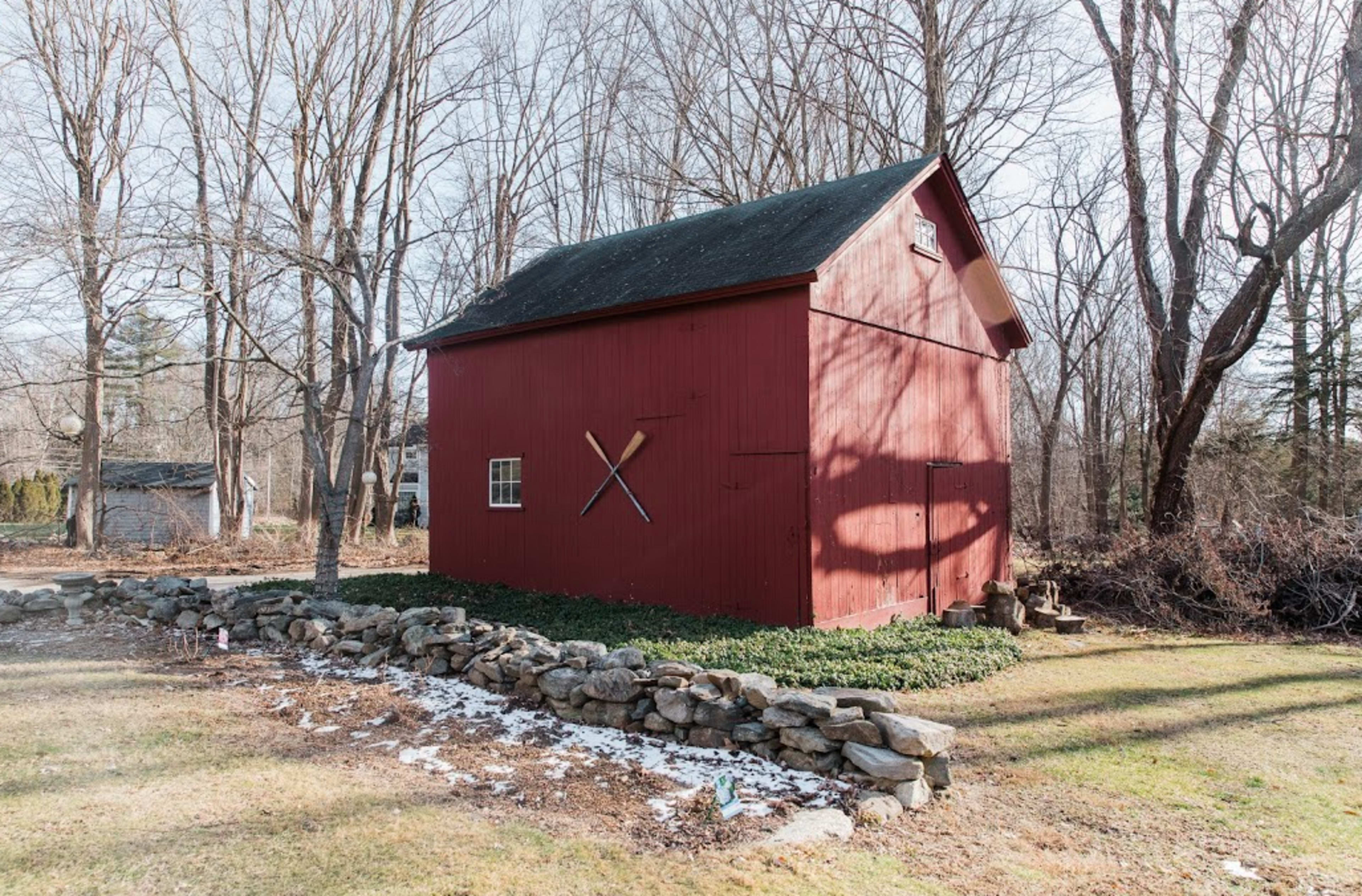 A red barn with two crossed wooden paddles is situated near a stone boundary in a wooded area.