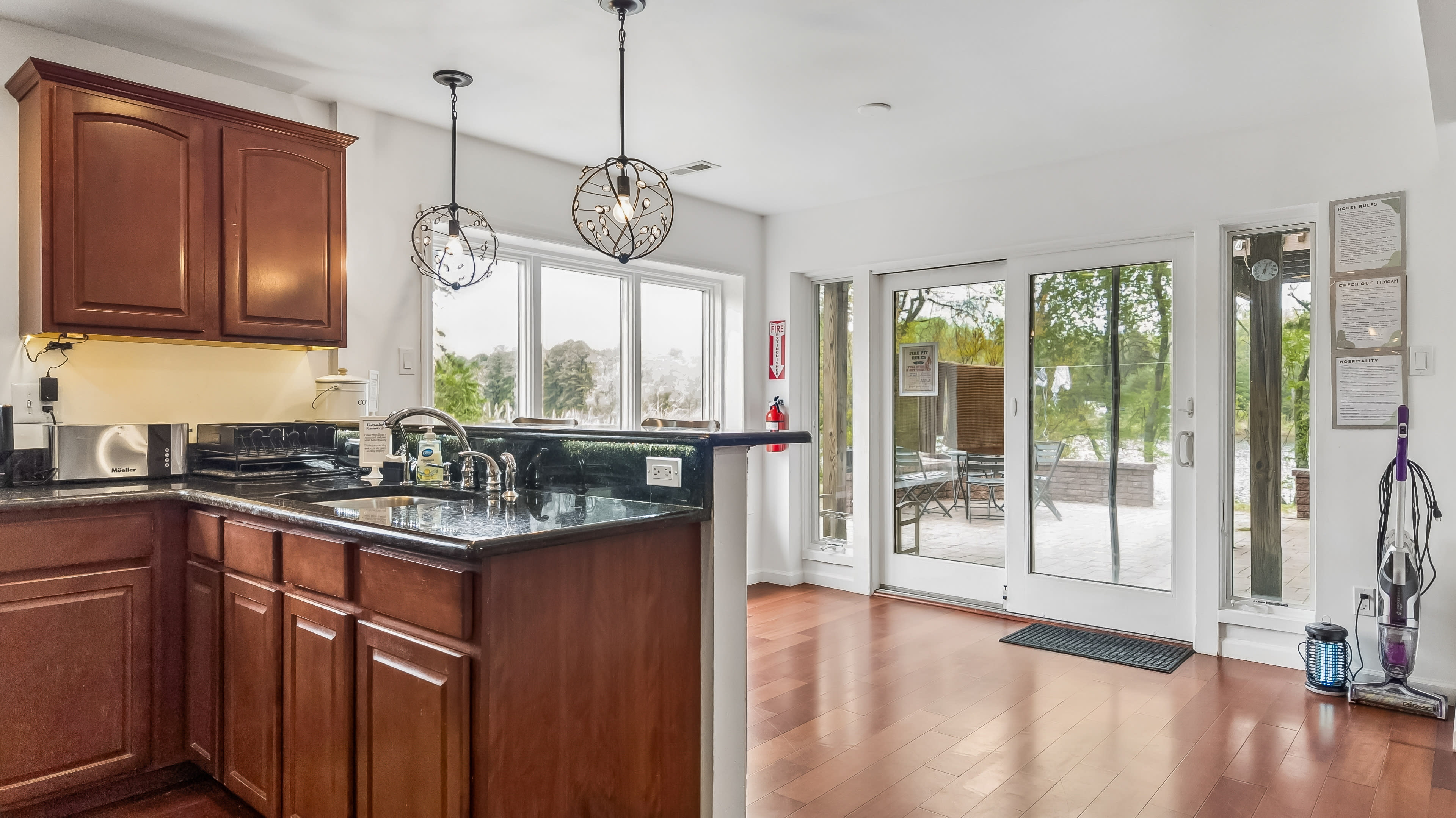 The image shows a modern kitchen with wooden cabinets and a countertop that opens into a sunlit dining area featuring sliding glass doors leading to an outdoor space.