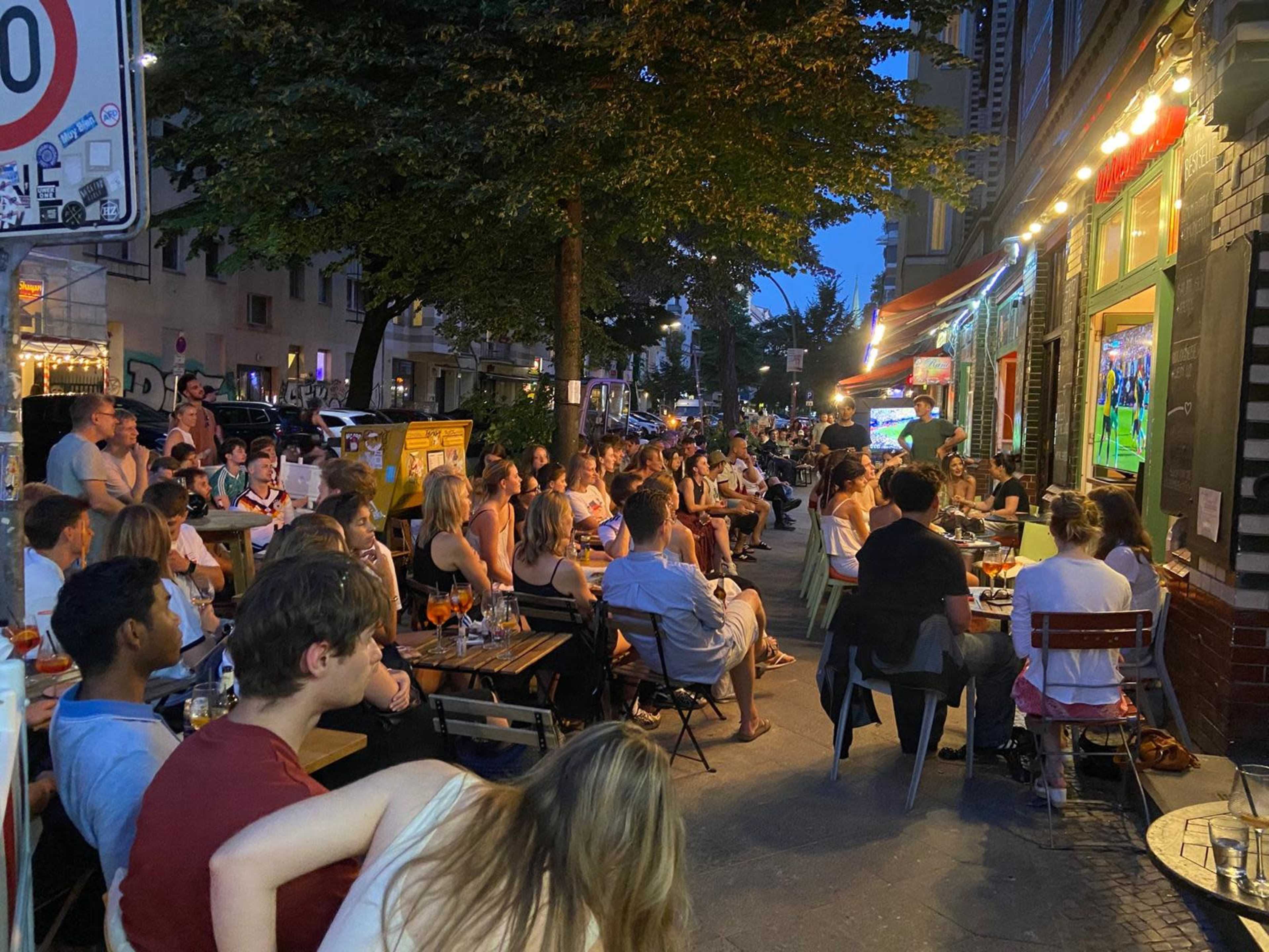 The scene shows a crowded outdoor café filled with people sitting at tables along a street in the evening.