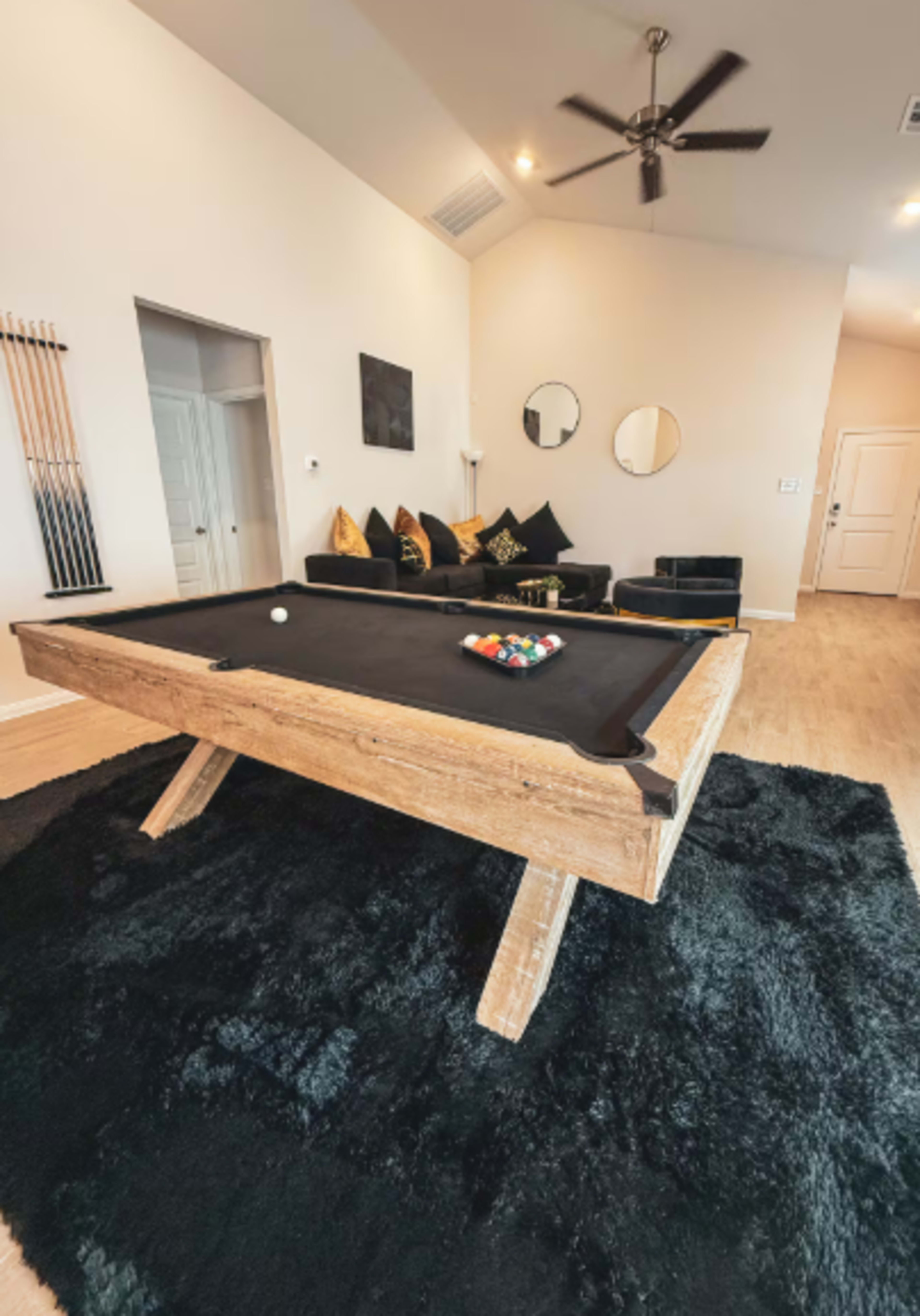 A wooden pool table with a black felt surface is positioned on a black rug in a brightly lit room featuring a couch and decorative mirrors.