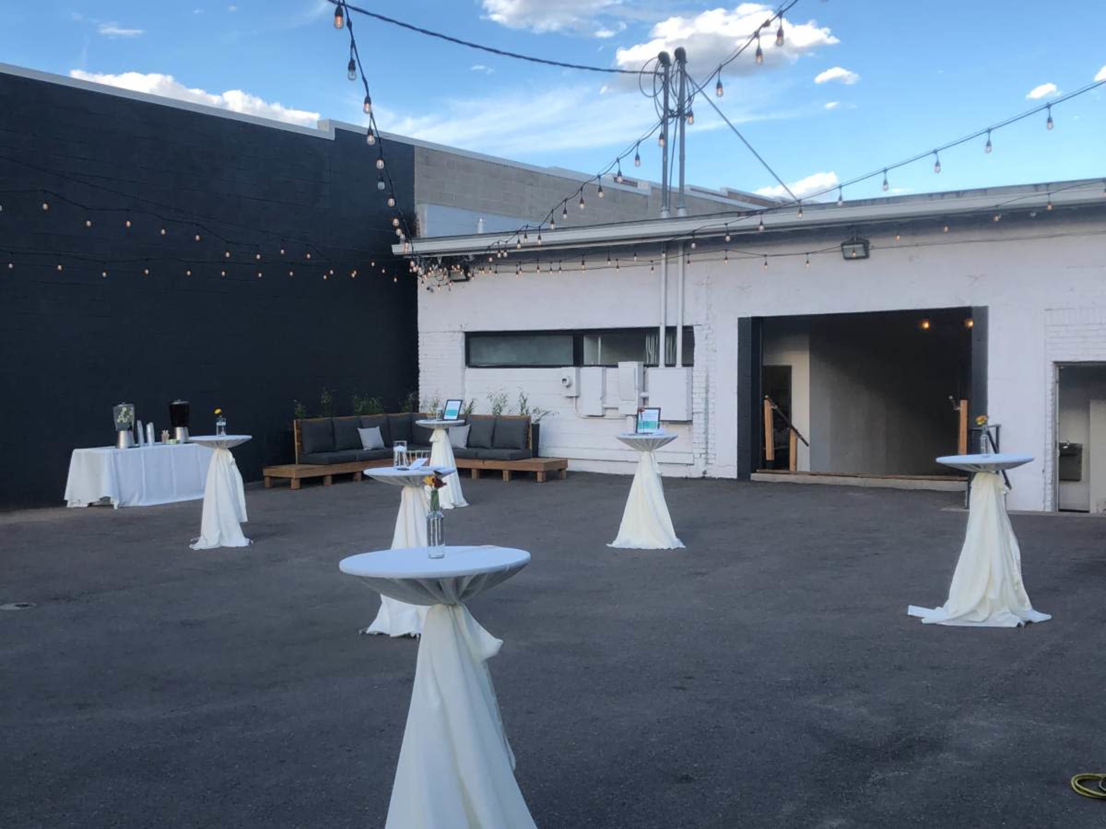 An outdoor event space with several cocktail tables covered in white tablecloths, string lights overhead, and a backdrop of a building.