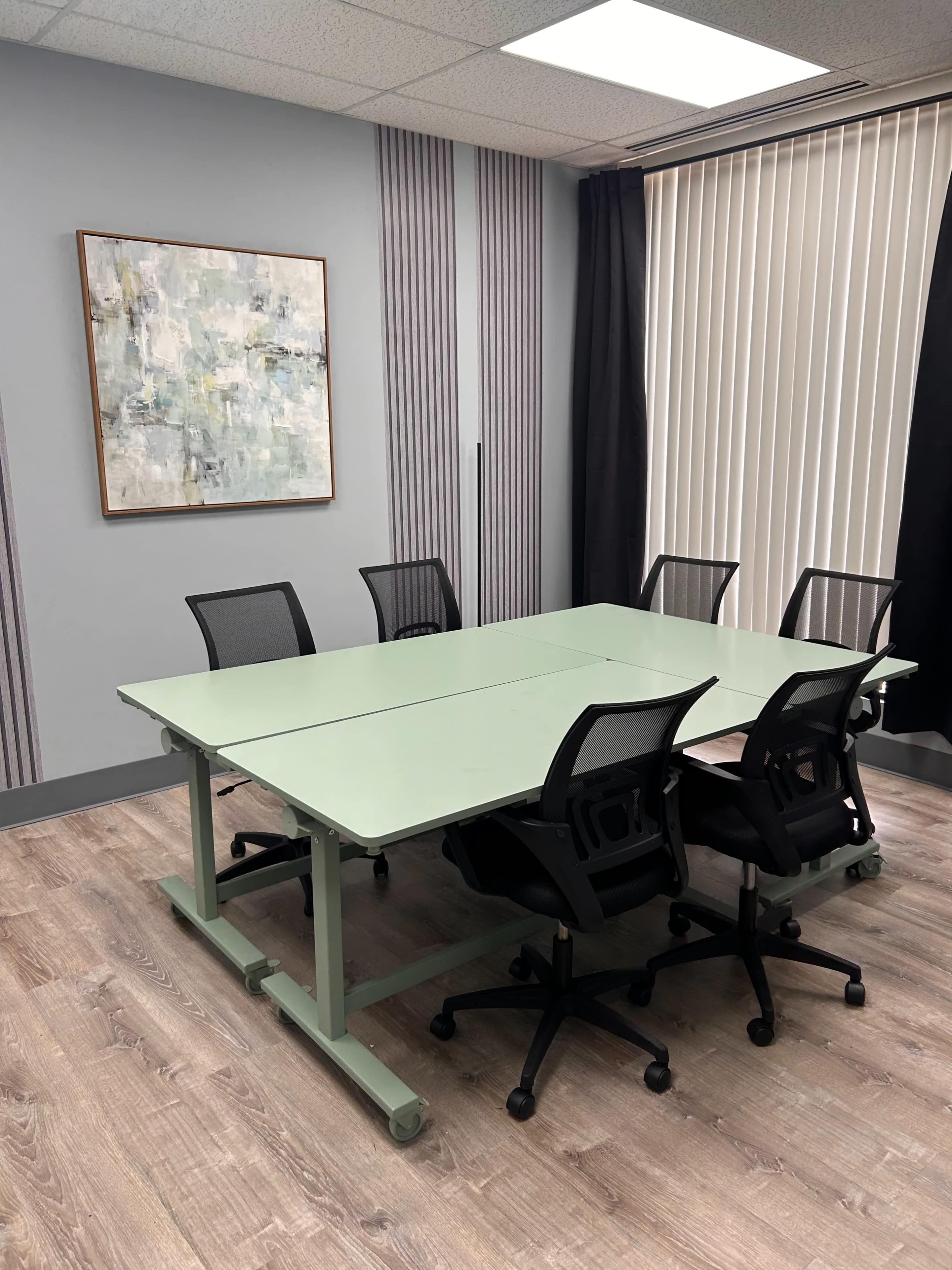 A light green conference table with six black mesh chairs is positioned in a well-lit meeting room featuring striped walls and a large window with blinds.