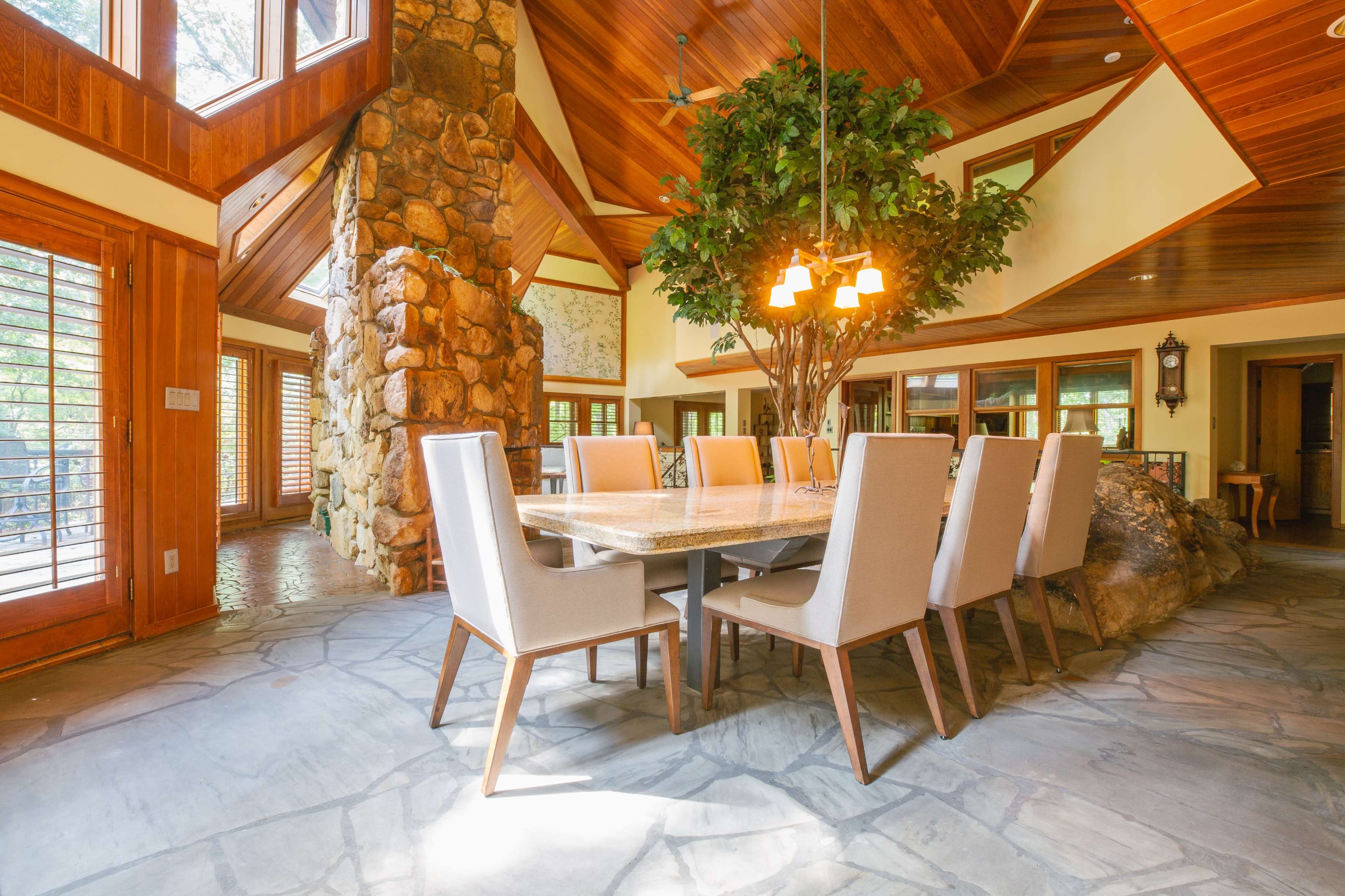 A dining area features a large table surrounded by upholstered chairs, with a stone wall and wooden beams in the background.