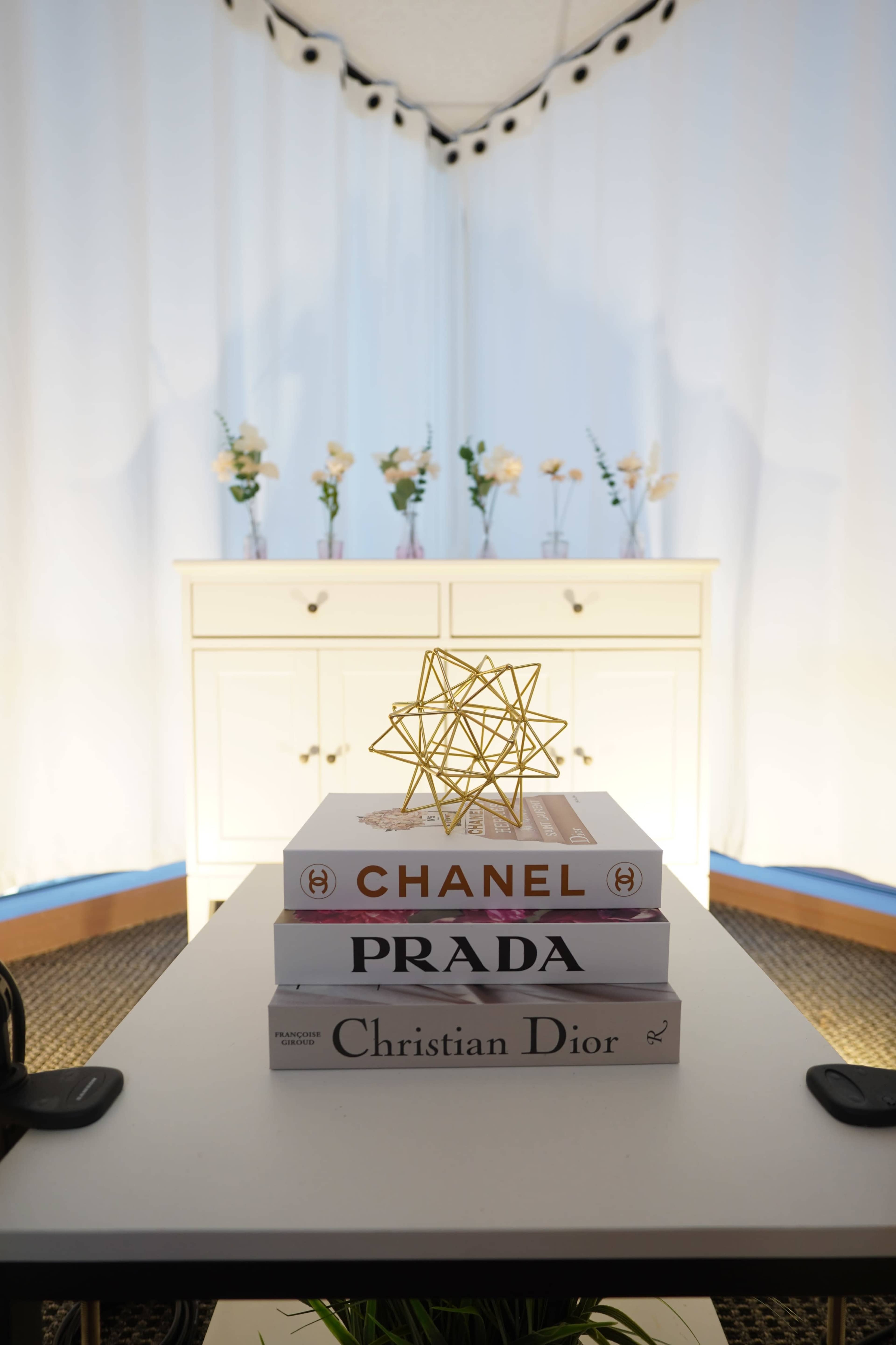A stack of fashion books topped with a geometric gold sculpture sits on a table in front of a white curtain backdrop adorned with small flower arrangements.