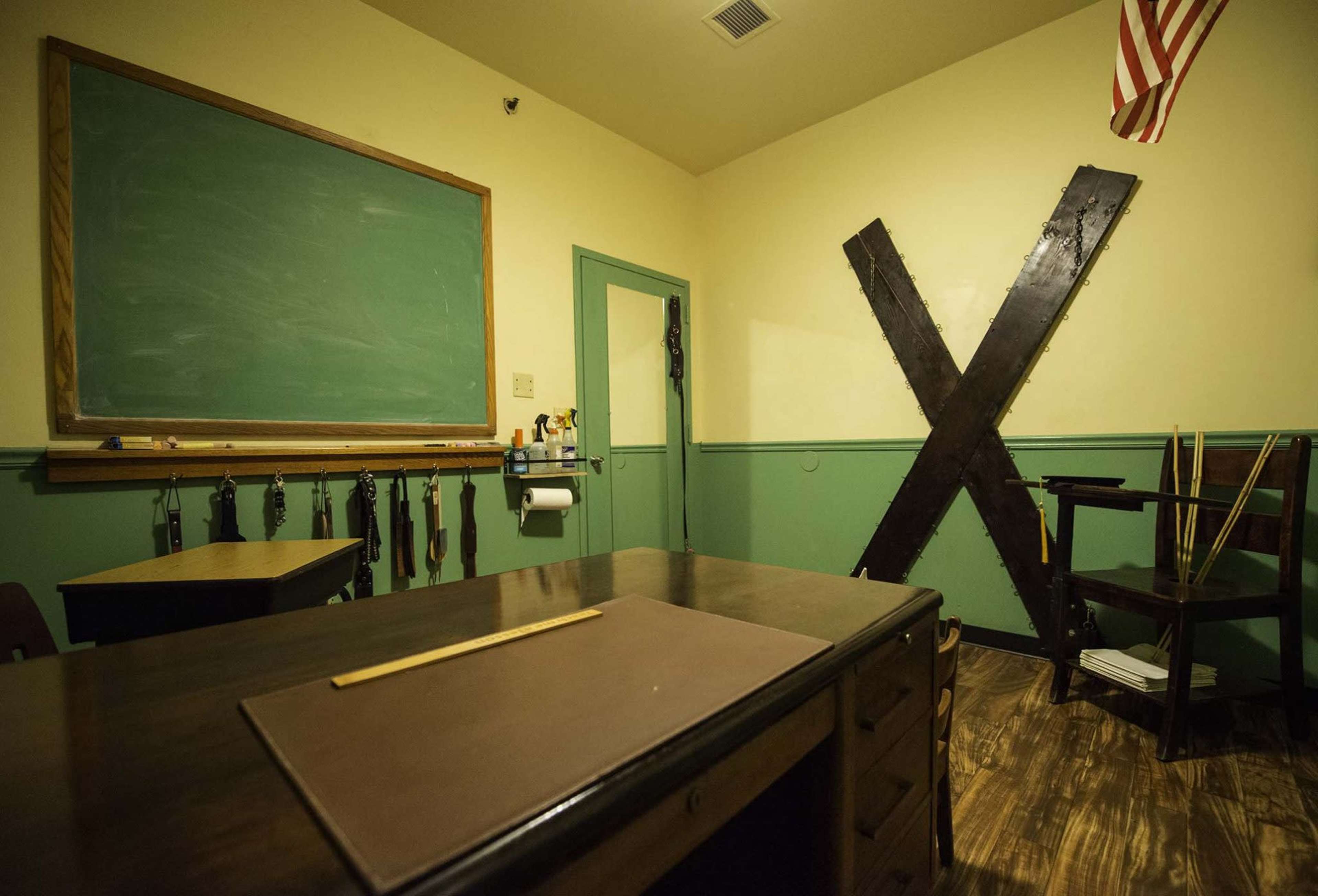 The image shows a small, empty classroom featuring a chalkboard, a large black wooden X on the wall, and a wooden desk with a writing pad.