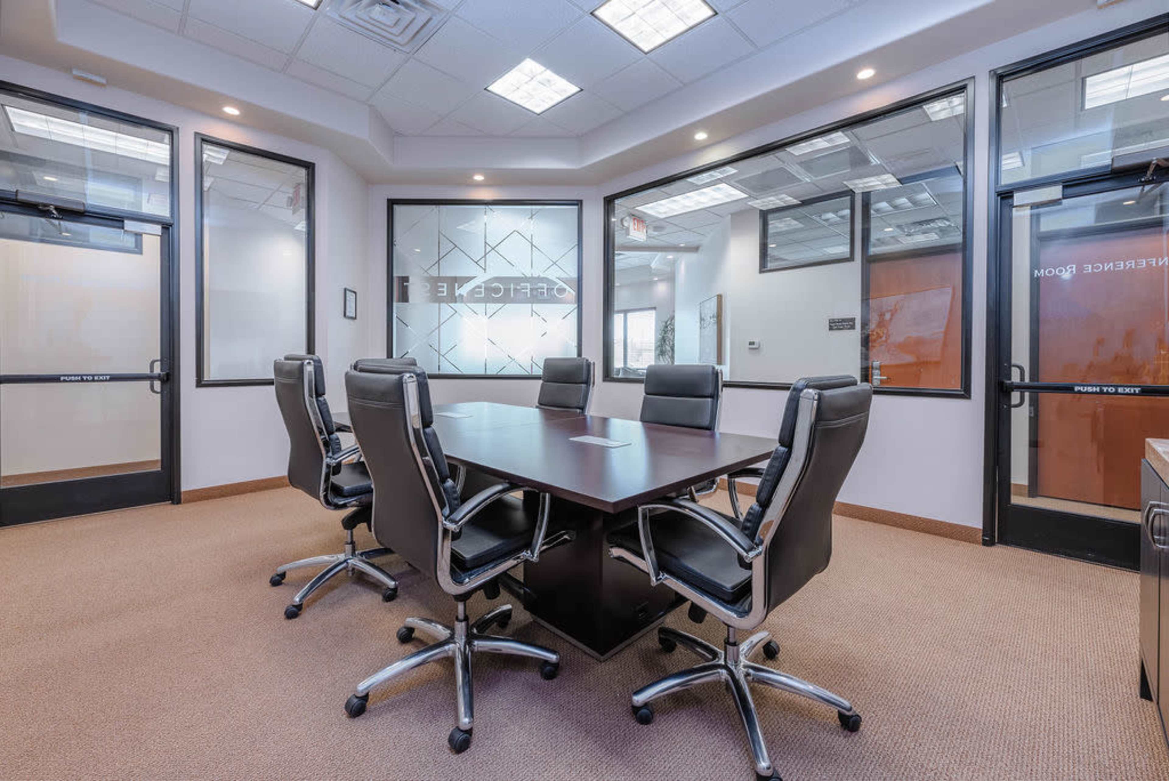 A modern conference room with a large wooden table surrounded by six black chairs, glass walls, and fluorescent lighting.