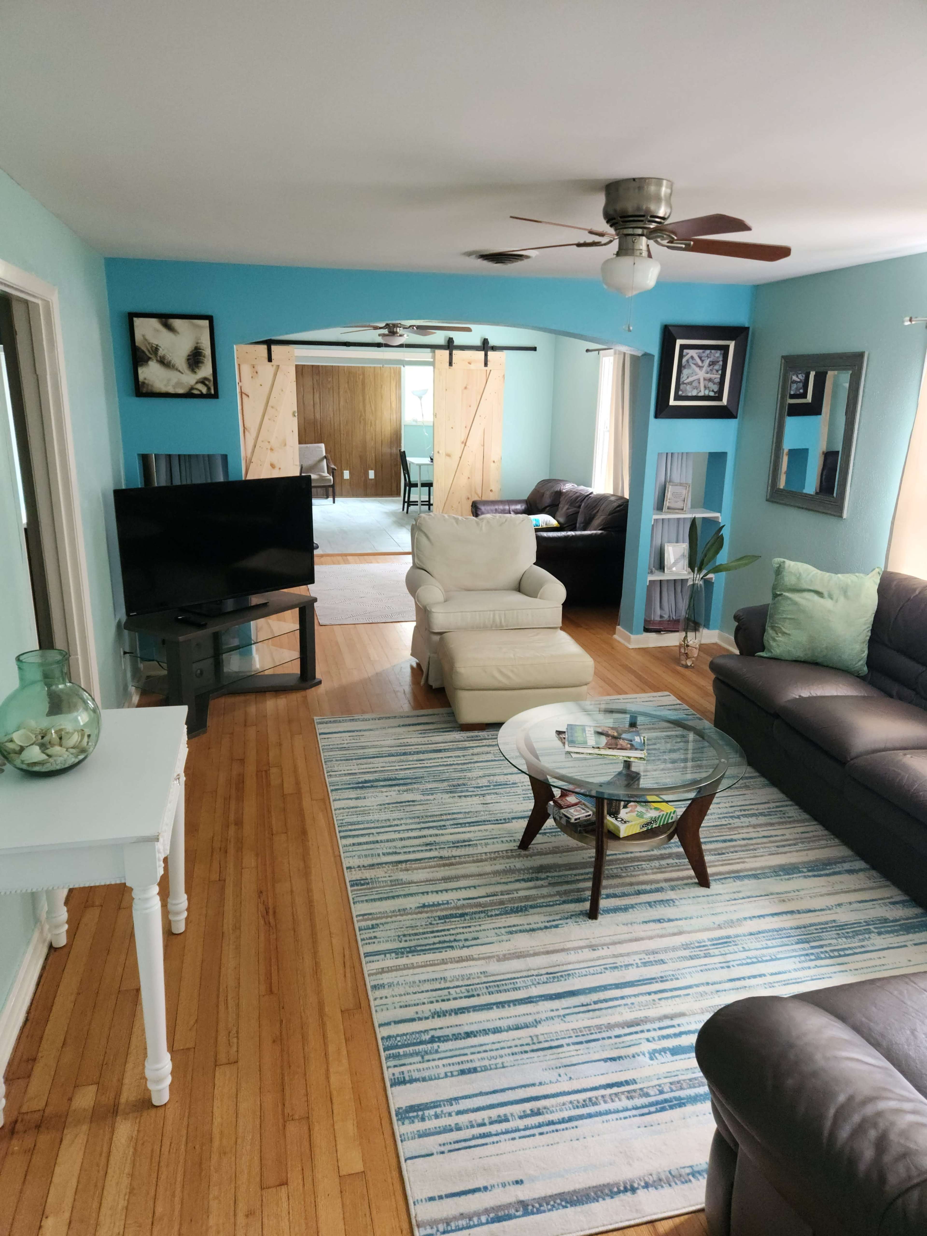 A living room features a light blue wall, a television, two couches, a coffee table, and a view of a room beyond with sliding barn doors.