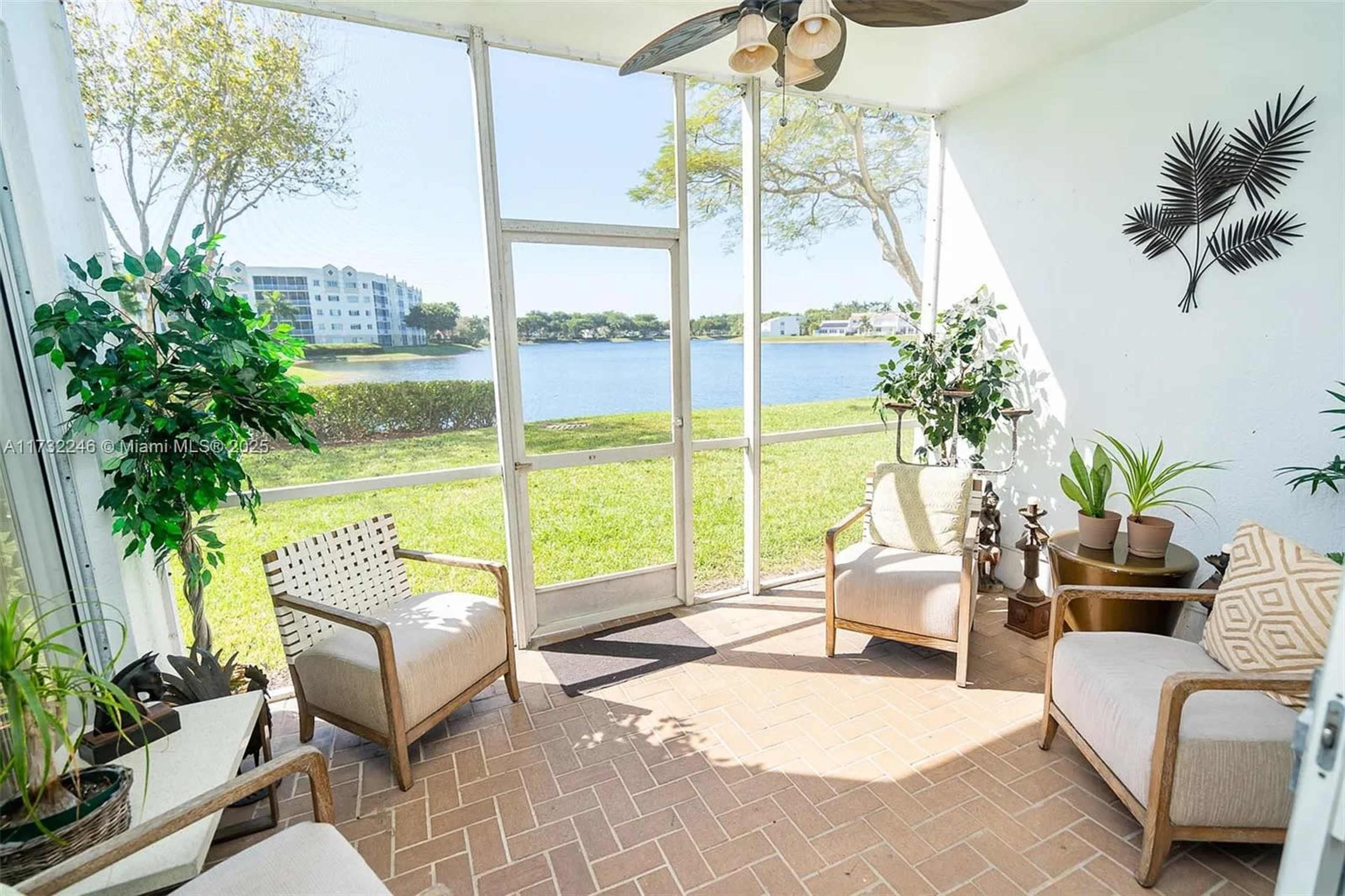 The image shows a sunlit screened porch with two chairs and decorative plants, overlooking a lake and greenery.