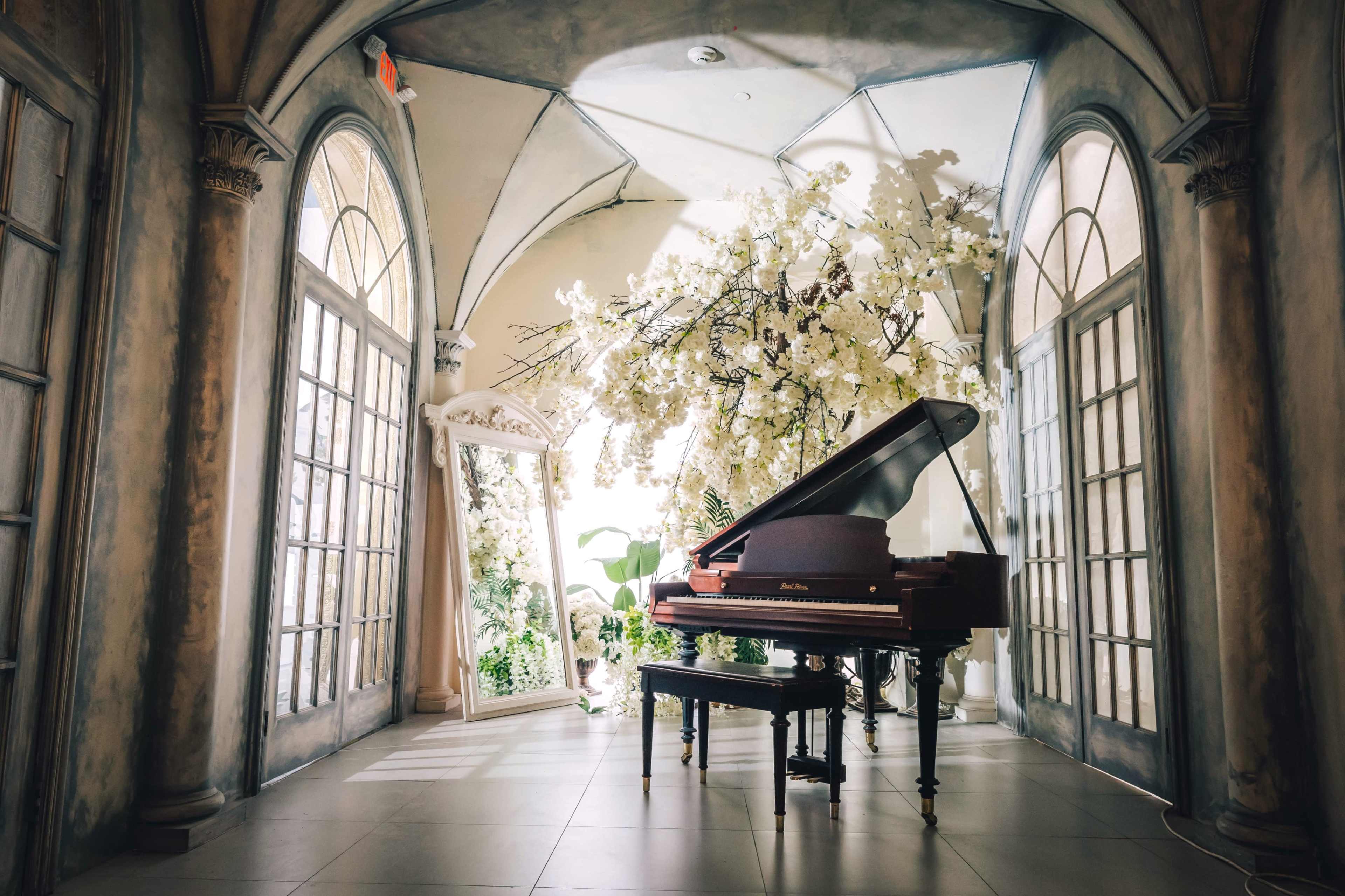 A grand piano is positioned in a bright room adorned with large windows and a mirror, surrounded by artificial cherry blossom branches.
