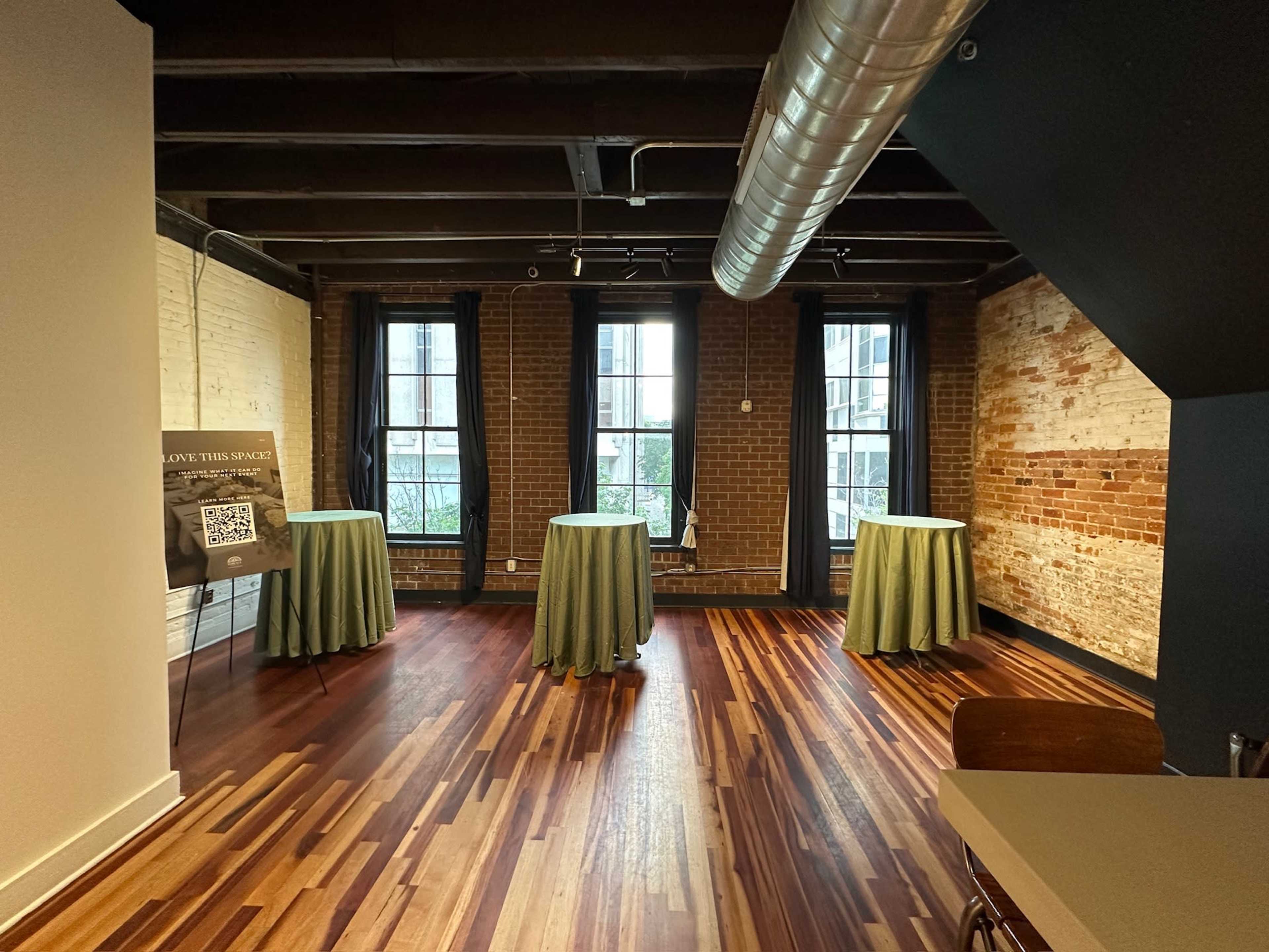 The image shows a spacious room with wooden floors, exposed brick walls, and three round tables covered with green tablecloths.