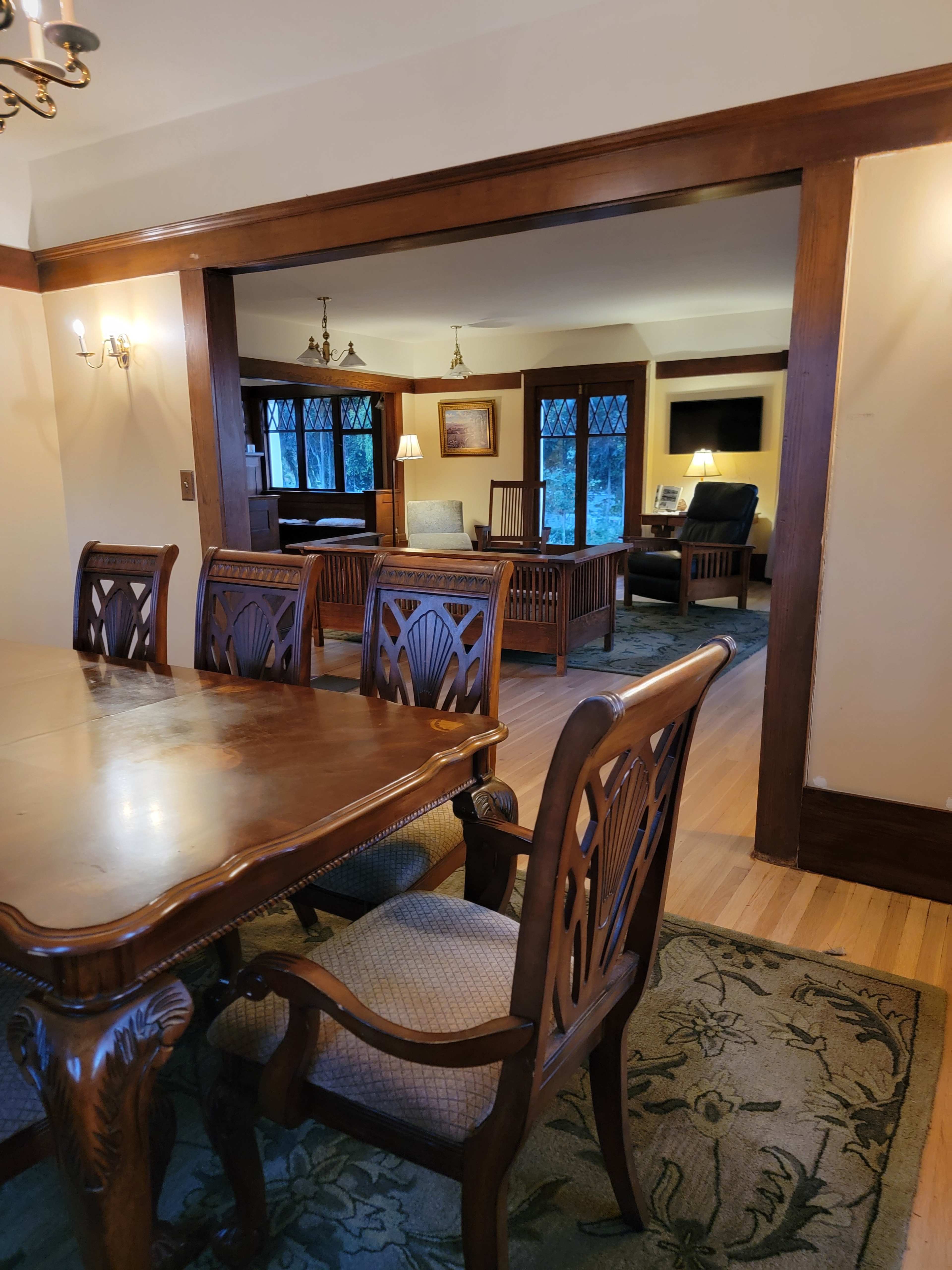 A wooden dining table with ornate chairs is positioned in the foreground, leading to a well-lit living room visible through an open archway.