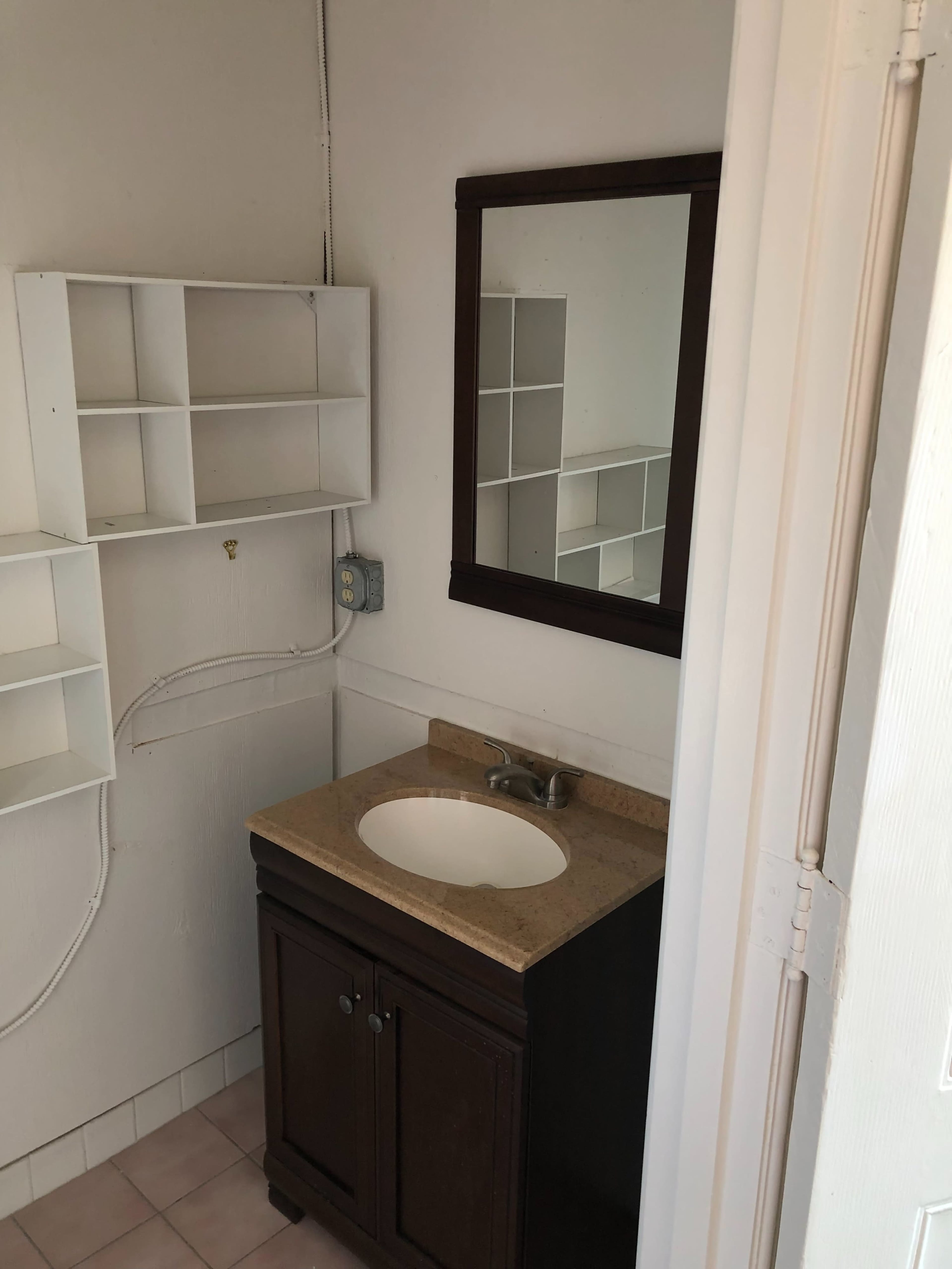 A simple bathroom with a wooden vanity, a sink, a mirror above it, and white shelving on the wall.