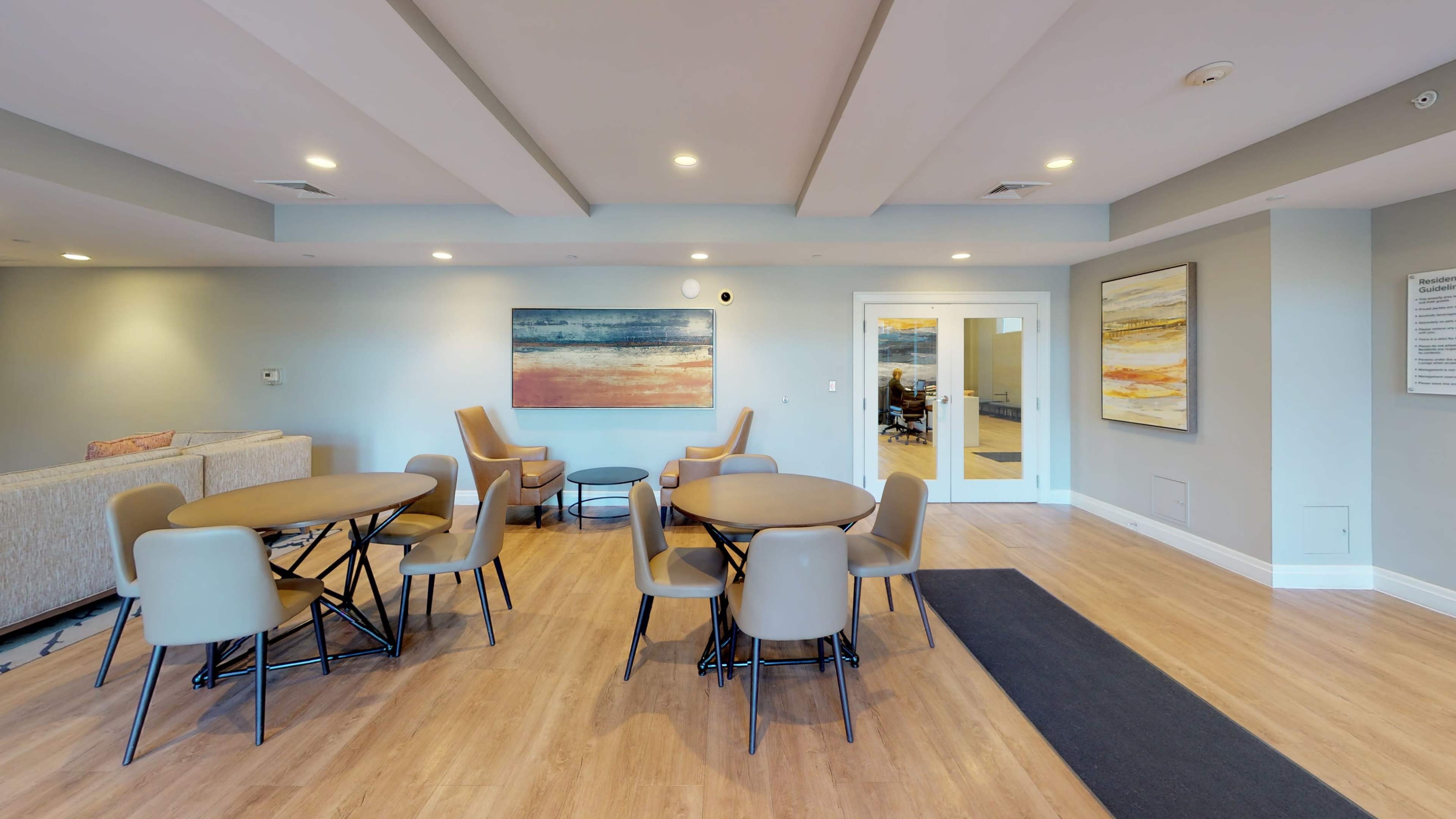 A modern lounge area featuring circular tables and chairs, with abstract artwork on the walls and a large window leading to another room.