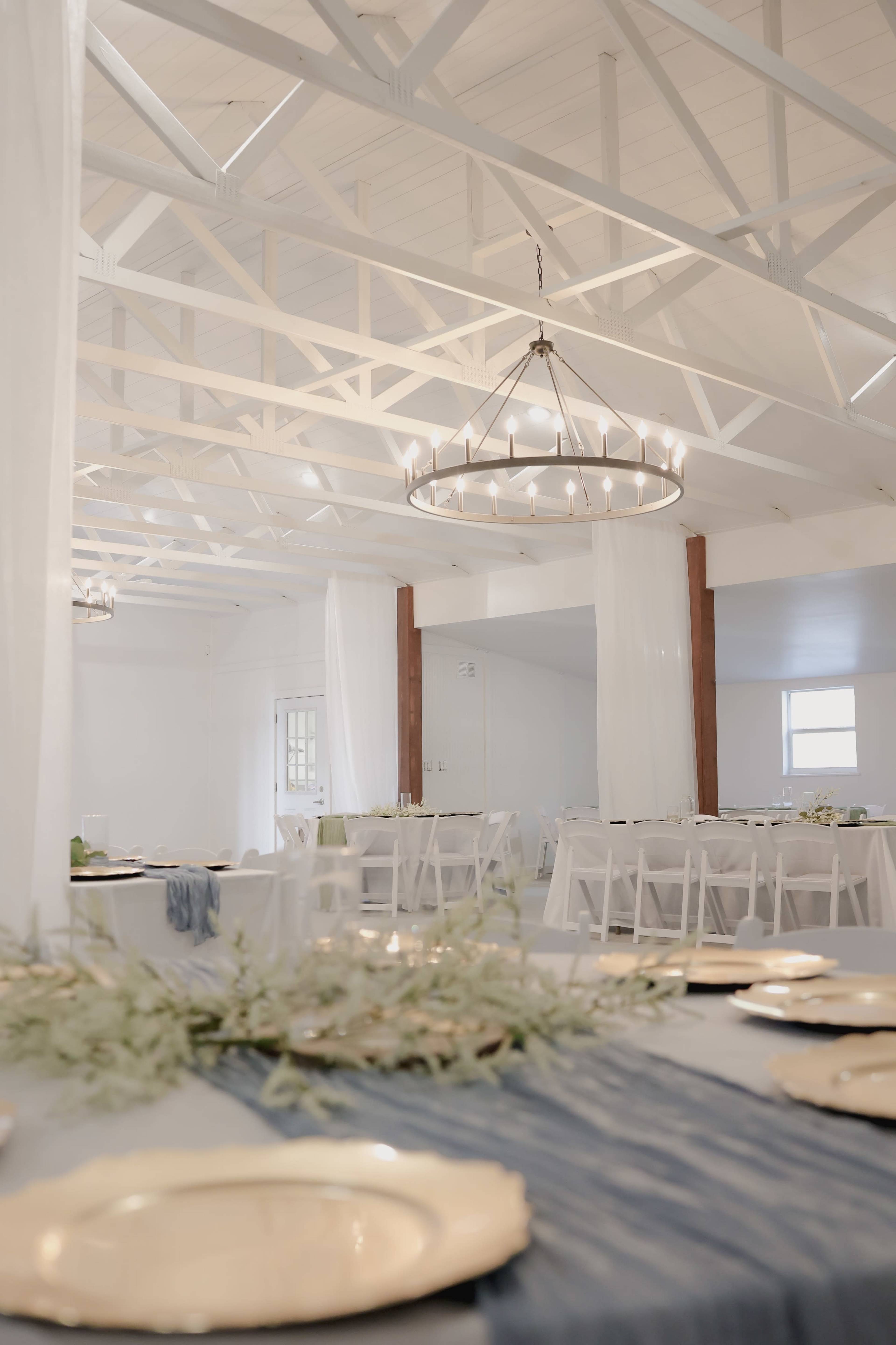 The image shows a spacious, elegantly arranged event venue with white walls and a light wood ceiling, featuring tables set for dining and decorative elements.