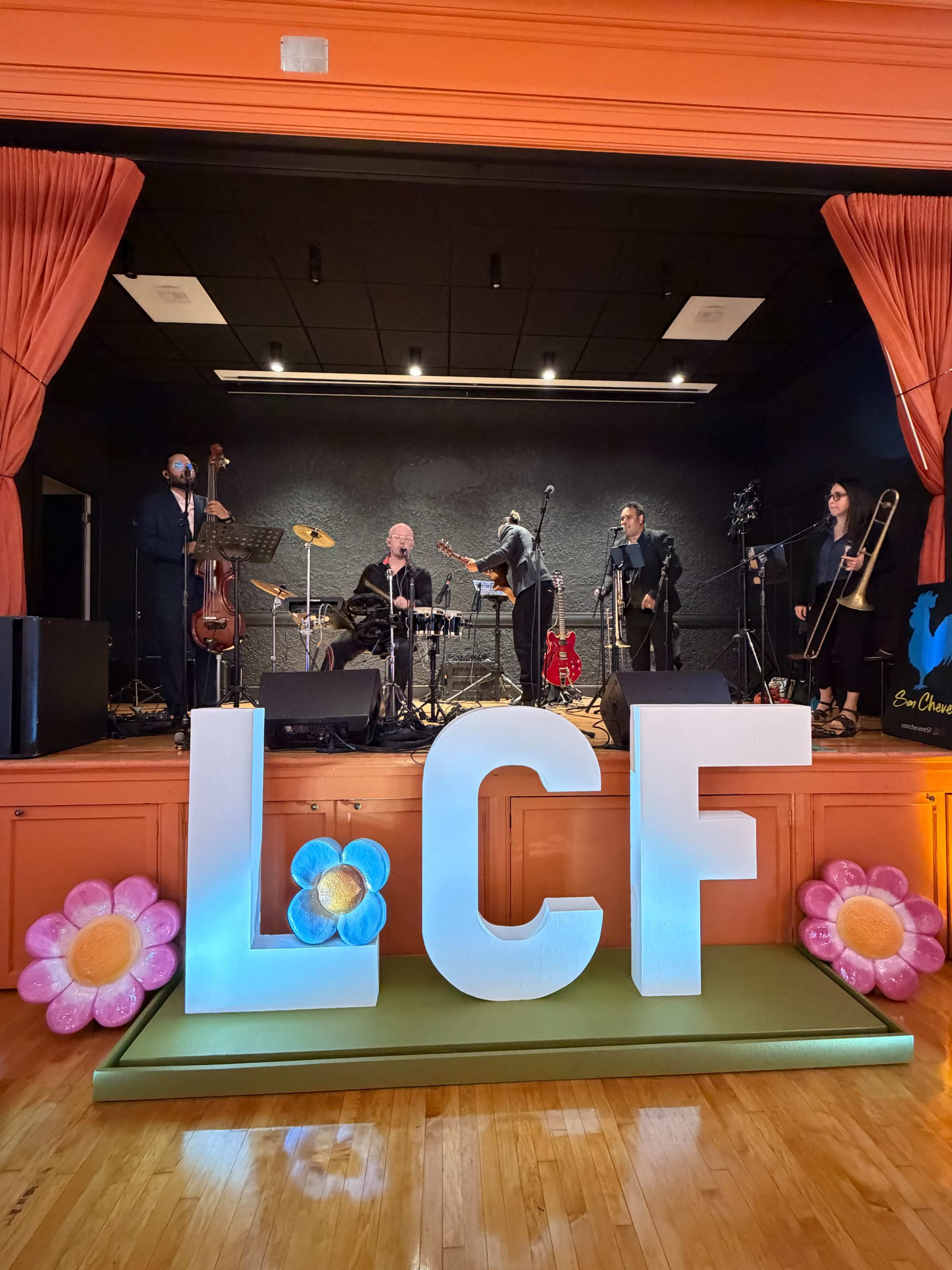 A live band performs on stage in front of large, colorful letters spelling "LCF," with decorative flowers in the foreground.