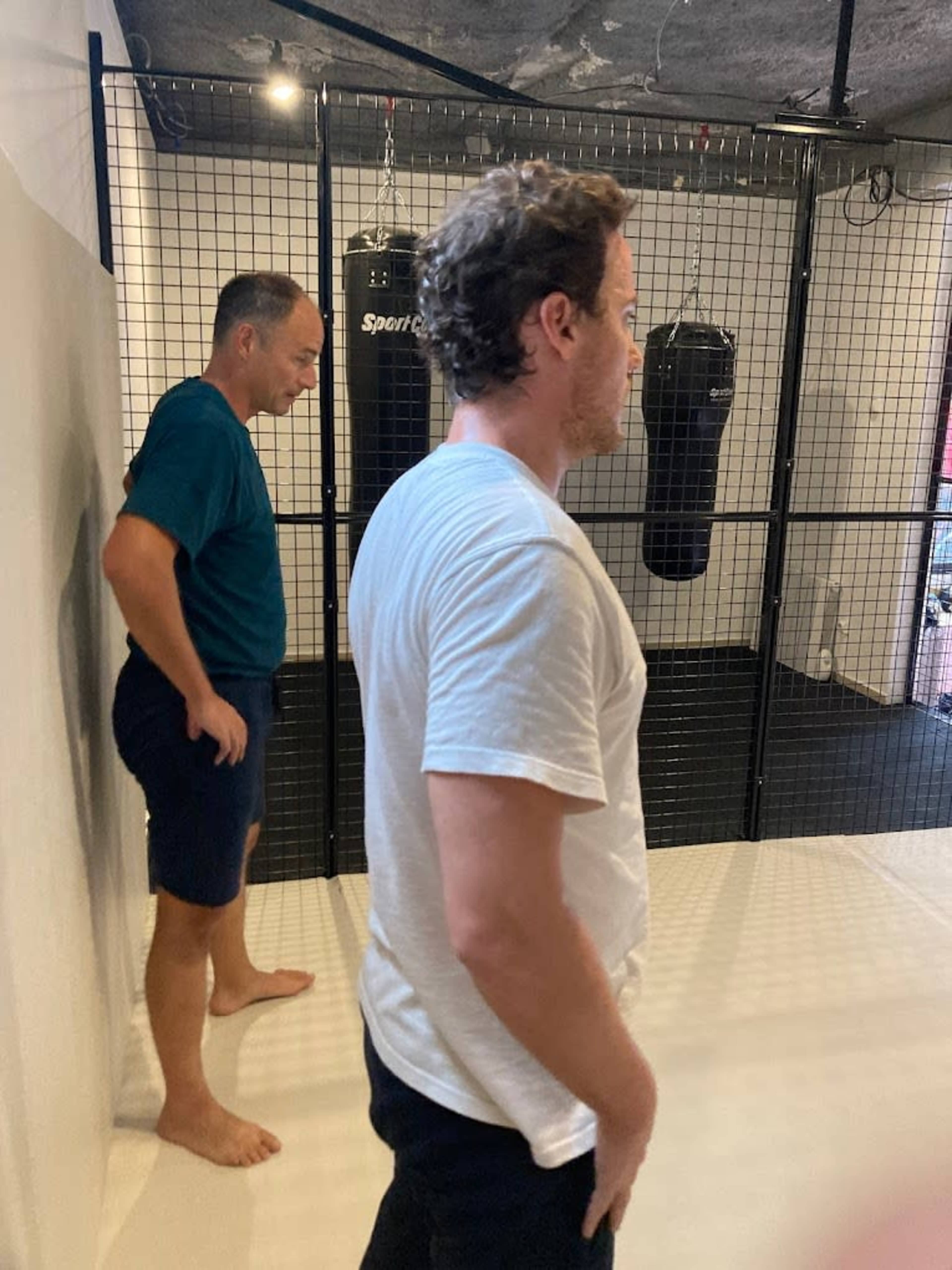 Two men stand inside a gym with boxing bags hanging in a caged area.