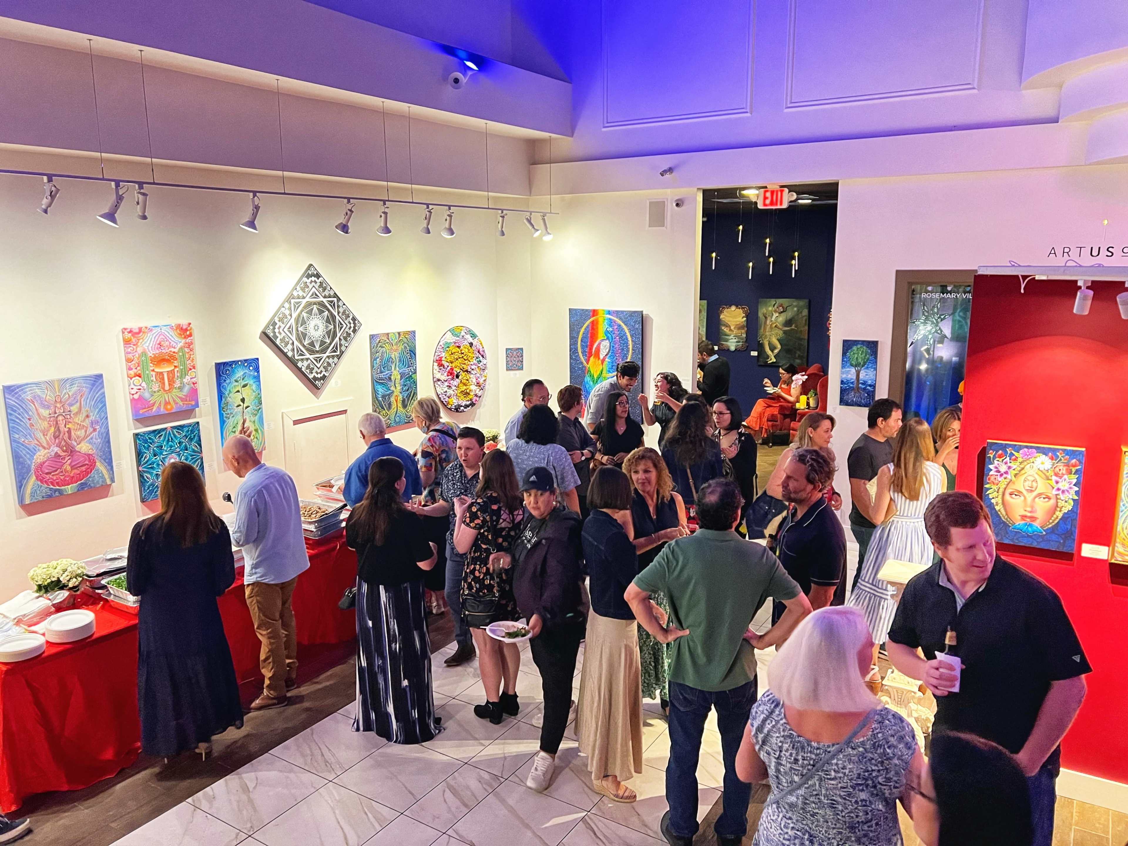 A gallery opening is taking place with attendees mingling, surrounded by colorful artwork displayed on the walls.