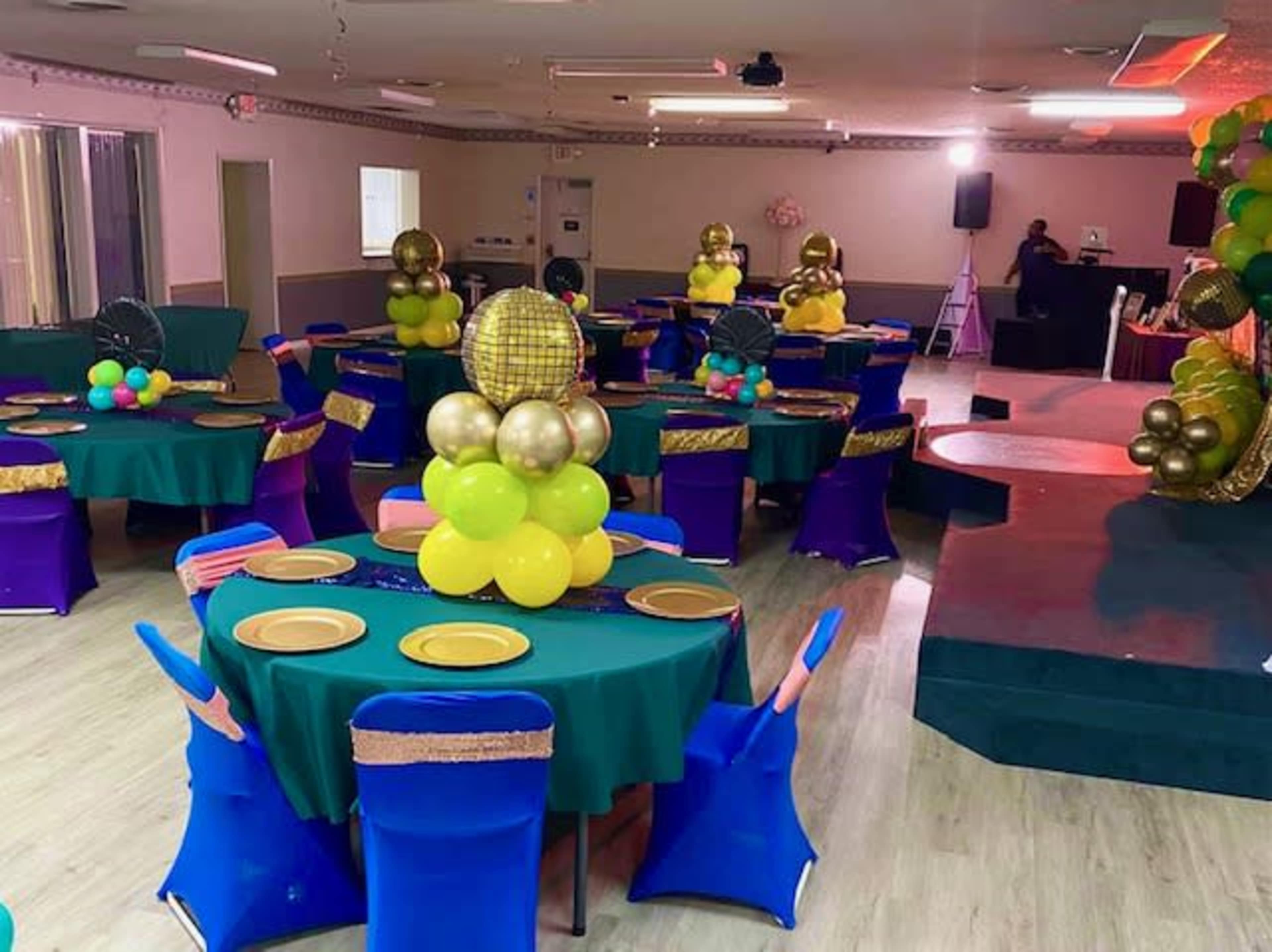 A decorated event space with round tables covered in green cloth, each featuring colorful centerpieces, and a small stage set for a performance.