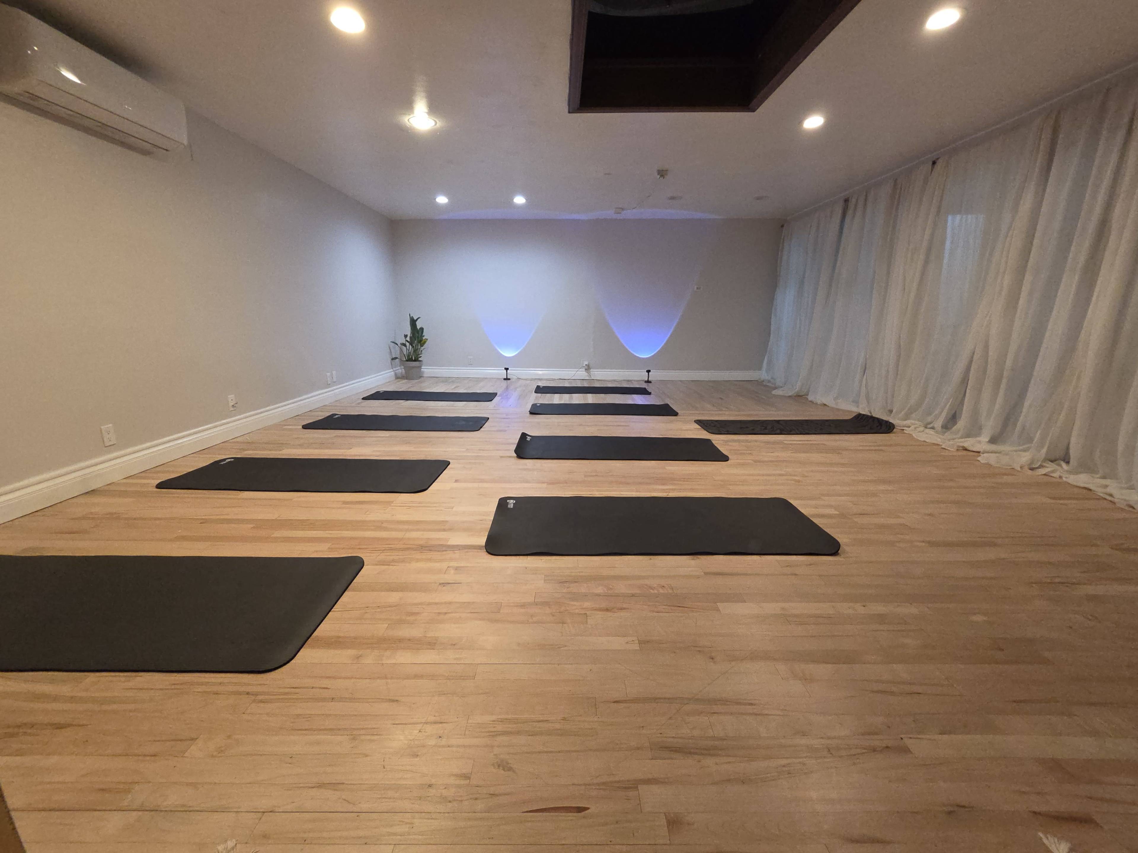 The image shows a spacious yoga studio with several exercise mats arranged on a wooden floor beneath soft lighting and sheer curtains.