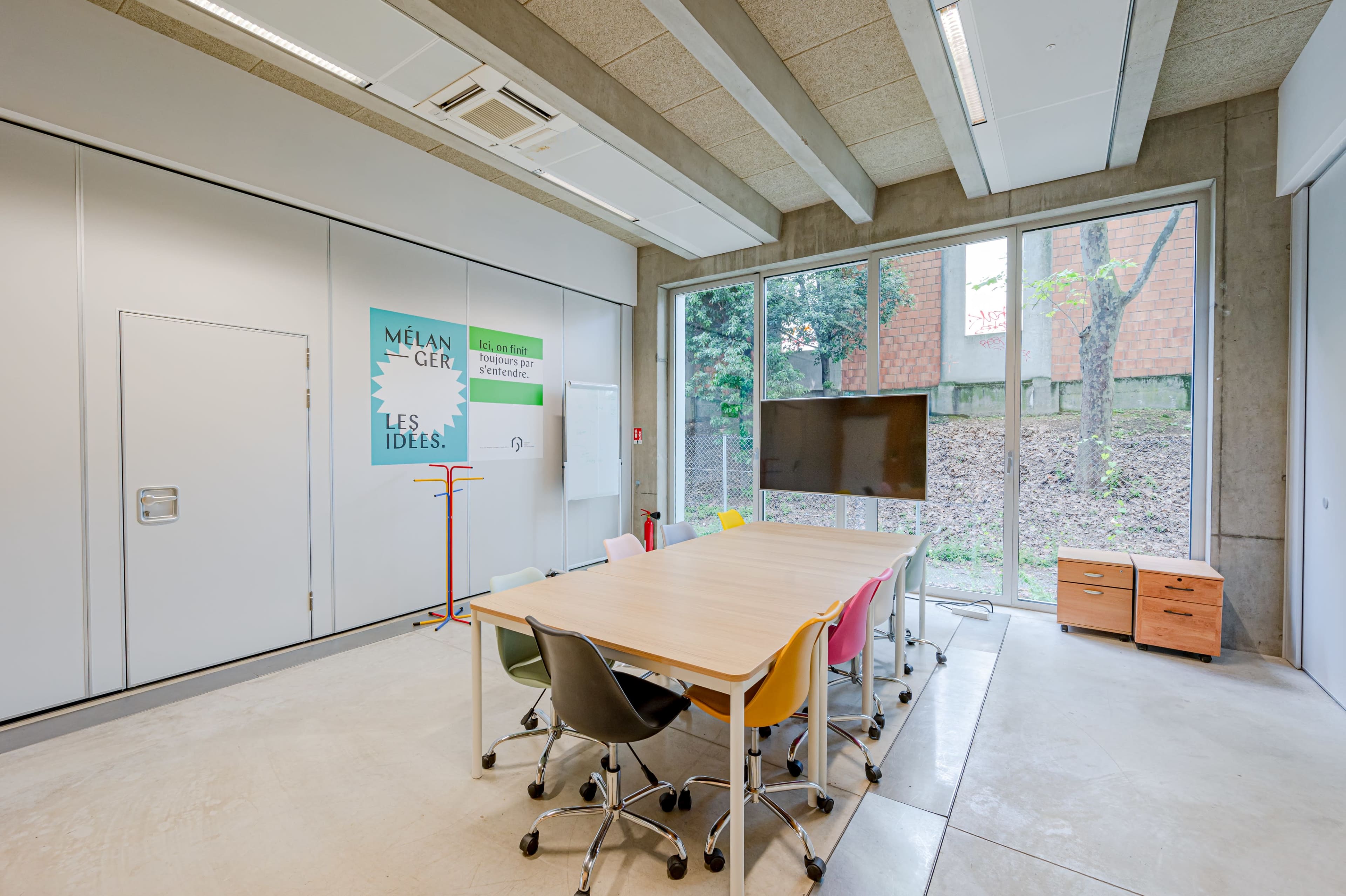 A modern conference room with a large table surrounded by colorful chairs, a wall-mounted screen, and windows overlooking a garden area.